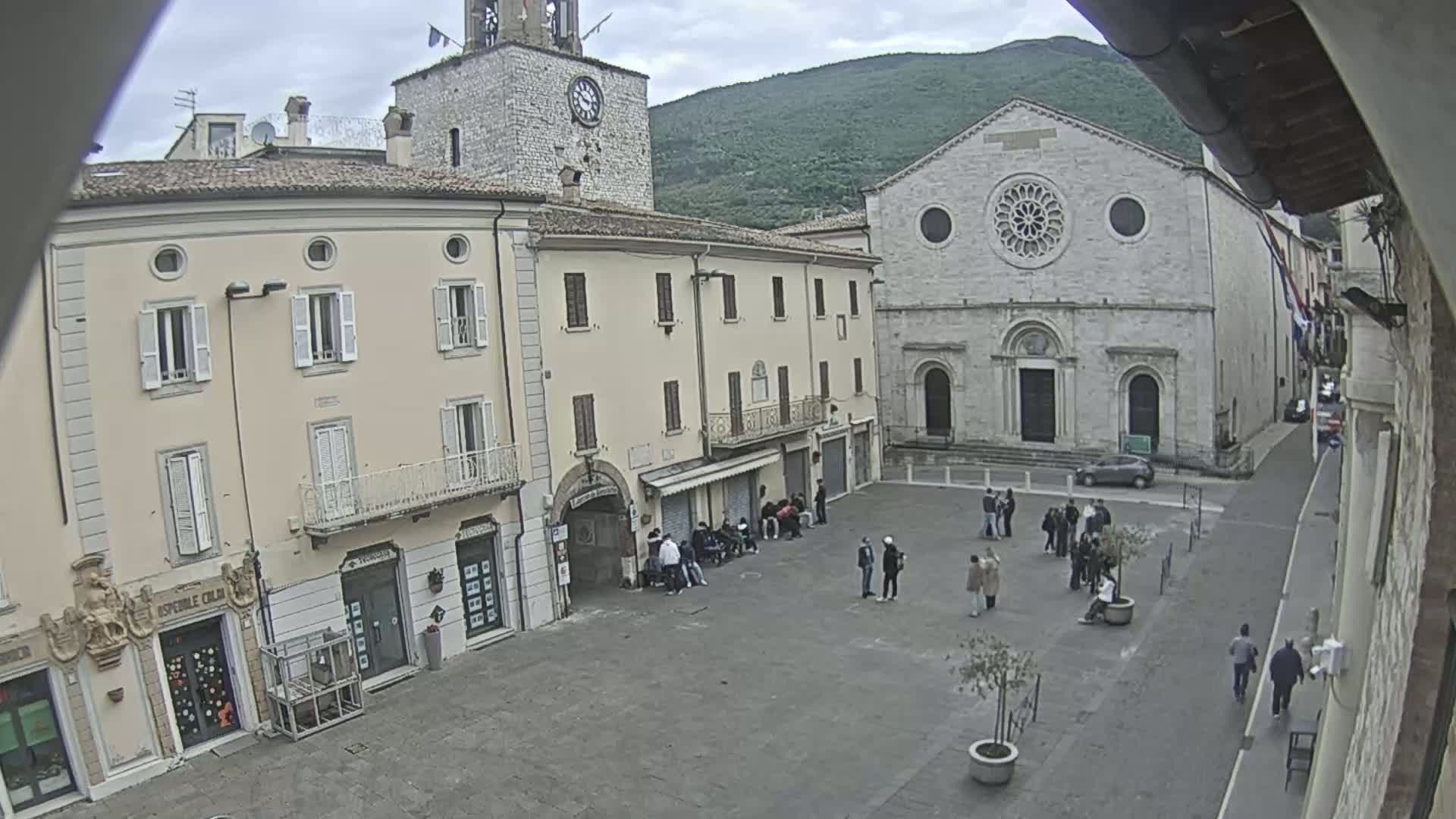 A town square with several people gathered, surrounded by buildings including a church and a clock tower, on an overcast day.
