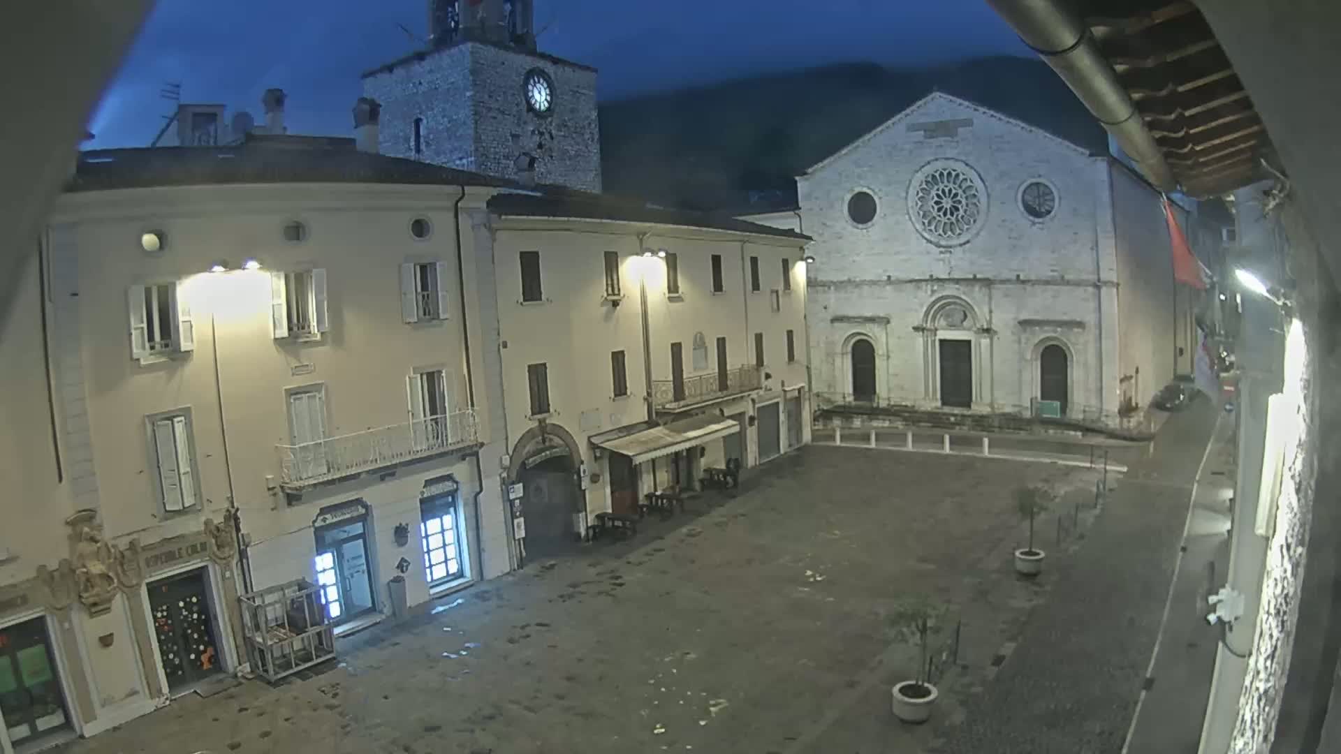 A nighttime view of a town square shows several buildings, including a clock tower and a church with a large rose window, under a dark, possibly overcast sky.