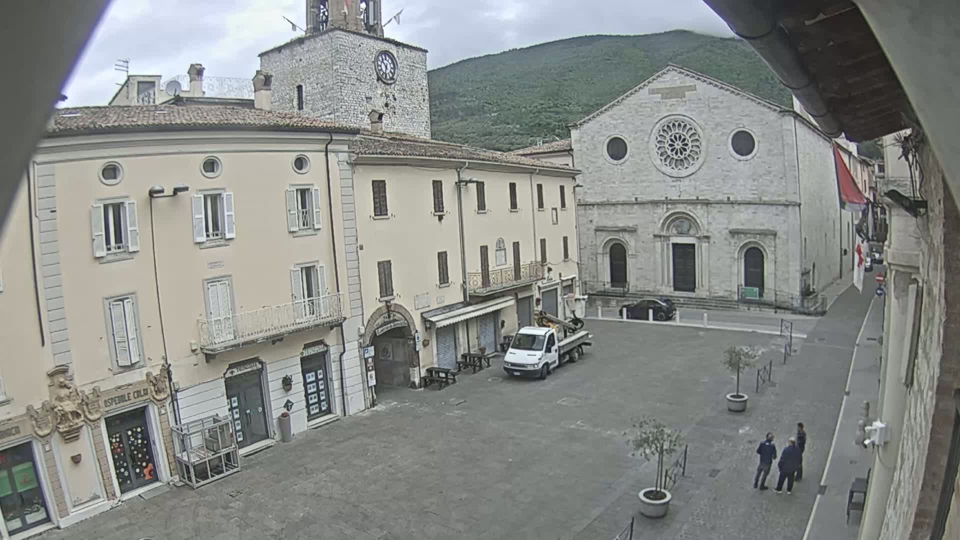 A mostly empty town square, under an overcast sky, is surrounded by buildings including a church with a rose window and a clock tower.