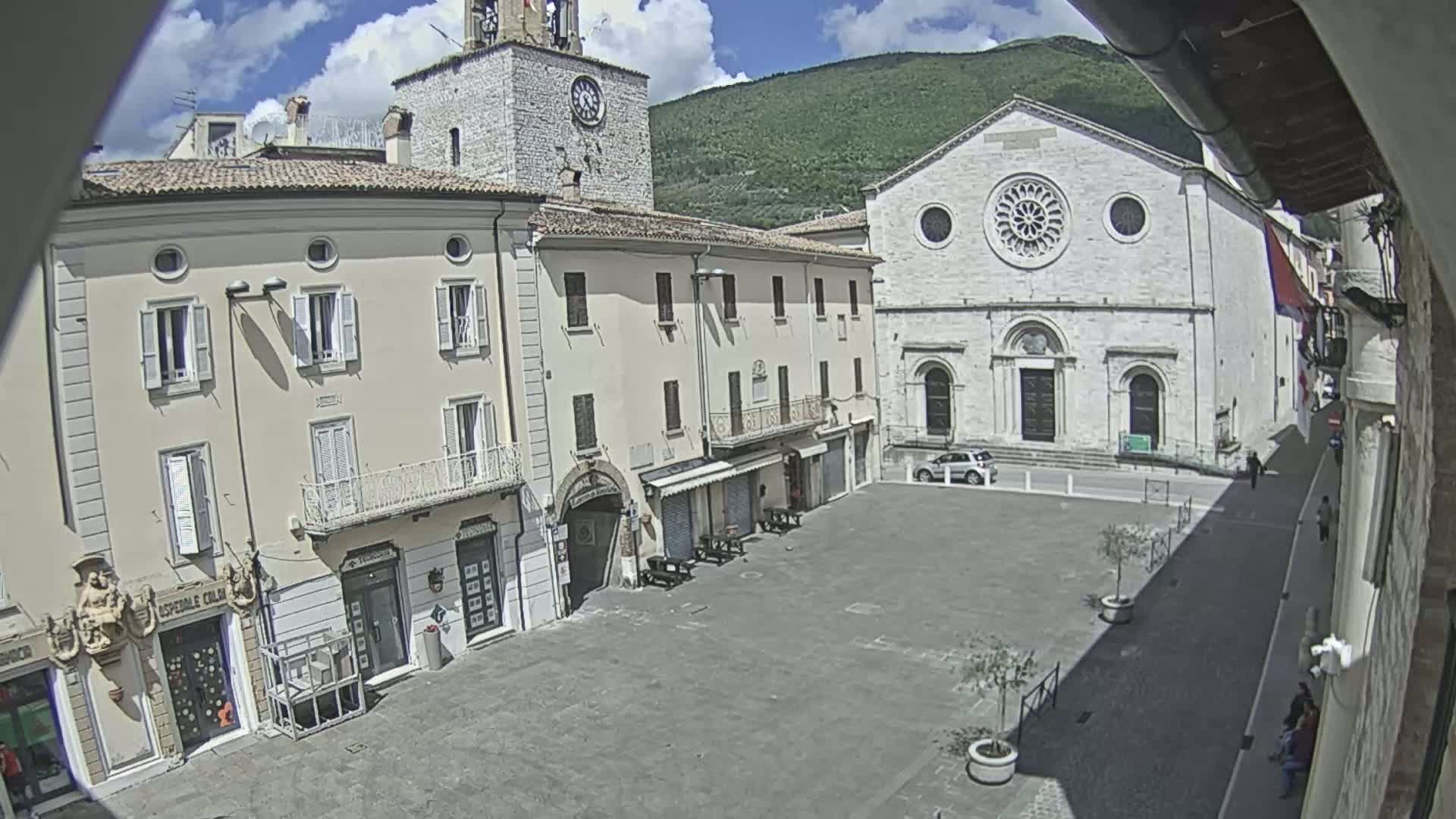 A mostly empty town square is surrounded by light-colored buildings, including a church with a rose window, under a partly sunny sky.