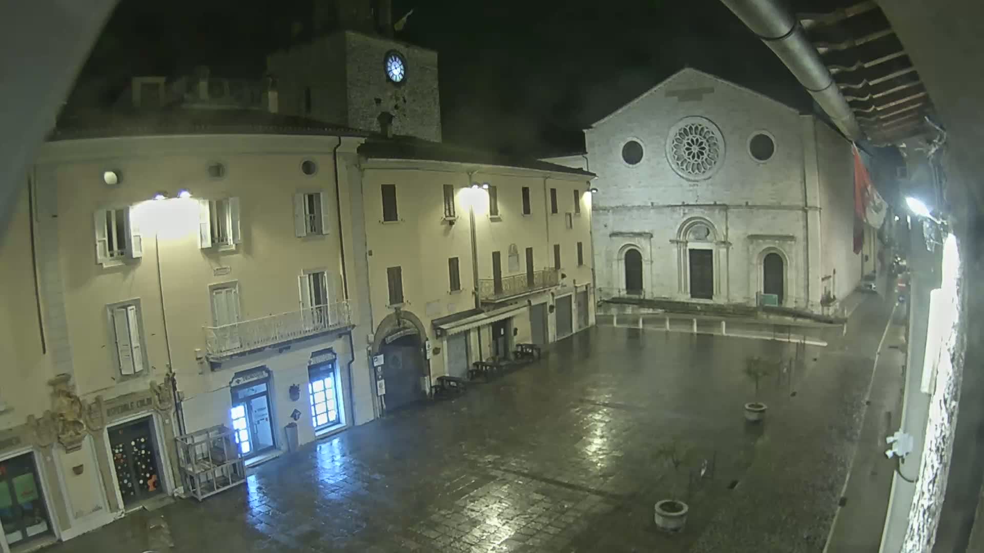 A rain-slicked town square at night is illuminated by streetlights, featuring buildings with classical architecture, including a church with a large rose window.