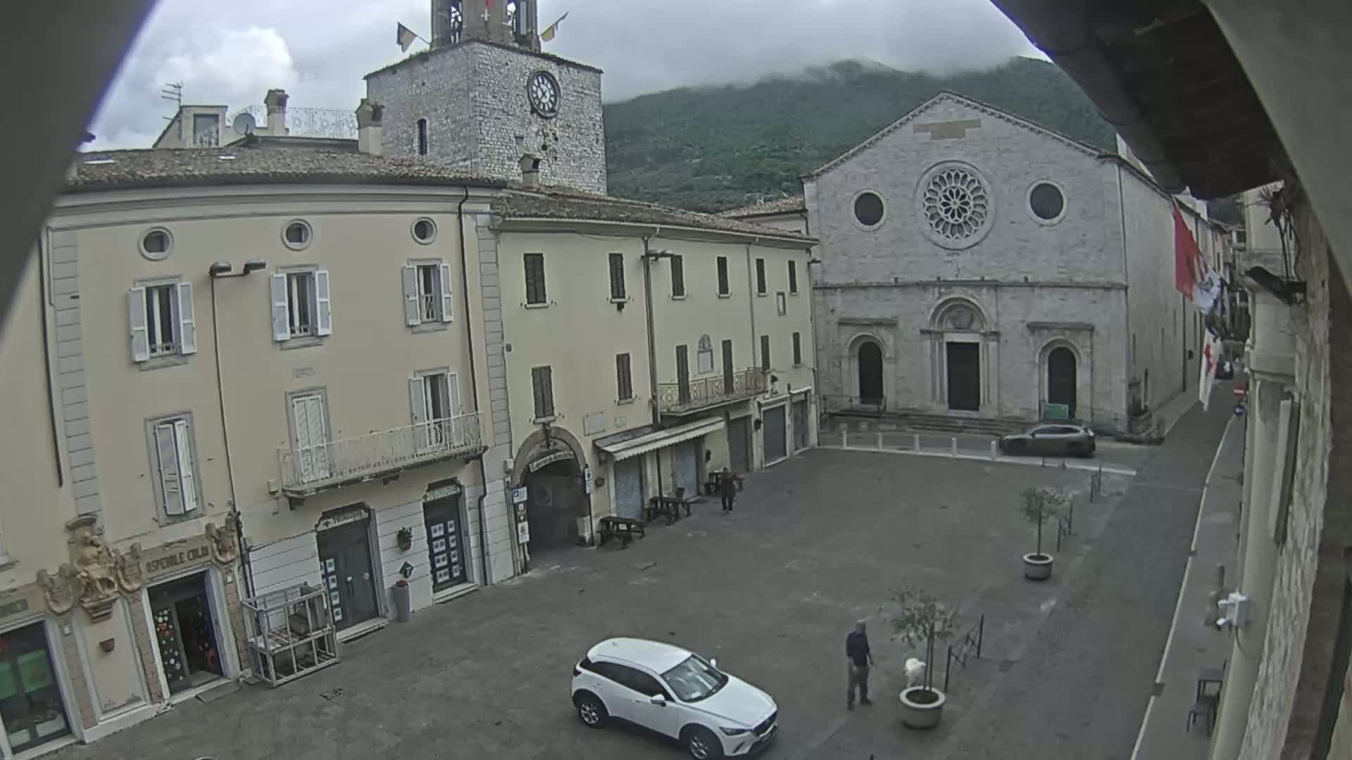 A mostly empty town square is surrounded by buildings, including a church with a rose window and a clock tower, under an overcast sky.