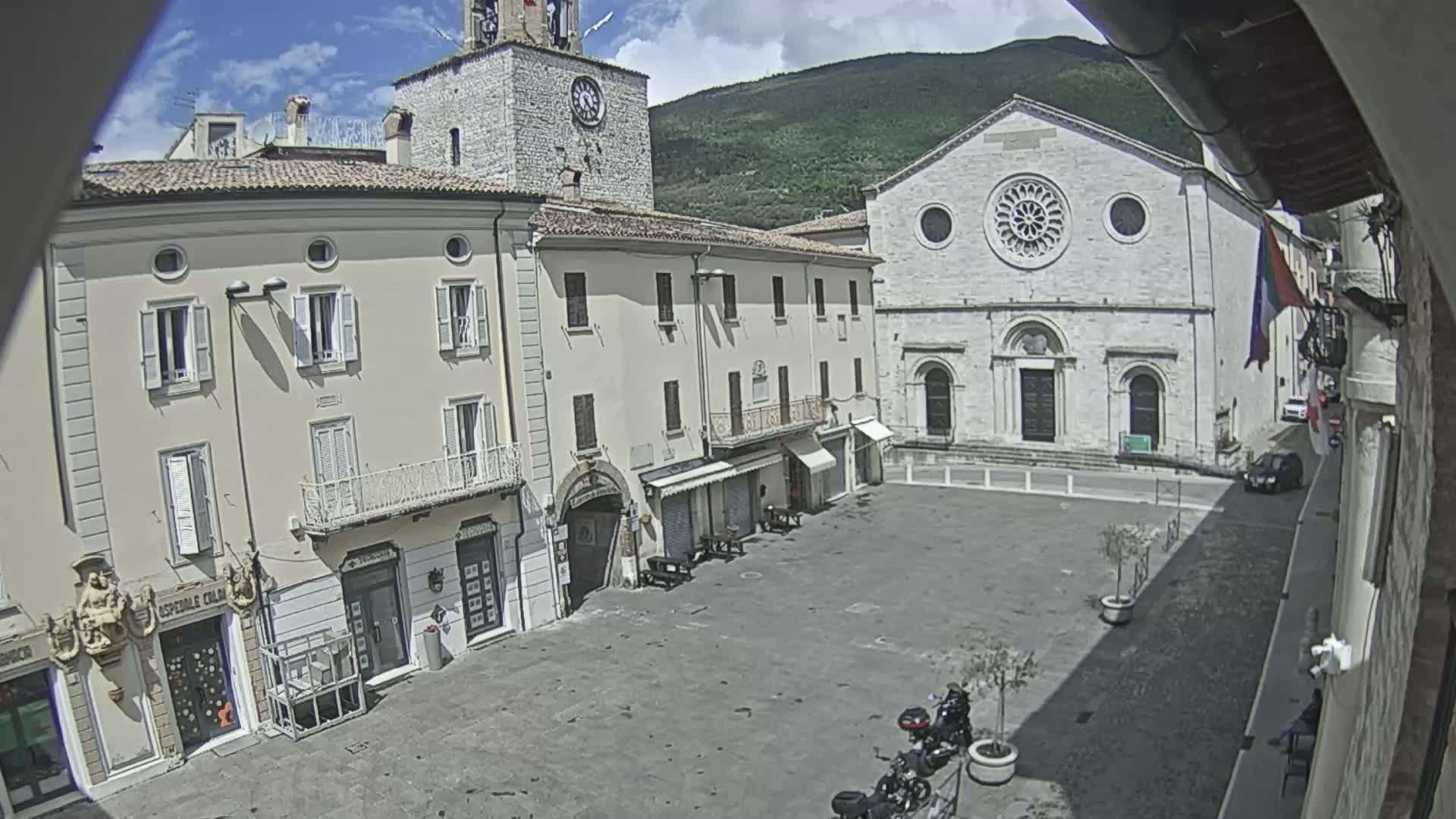 A sunny day reveals a town square surrounded by light-colored buildings, including a church with a rose window, and a clock tower, with a few motorcycles parked nearby.