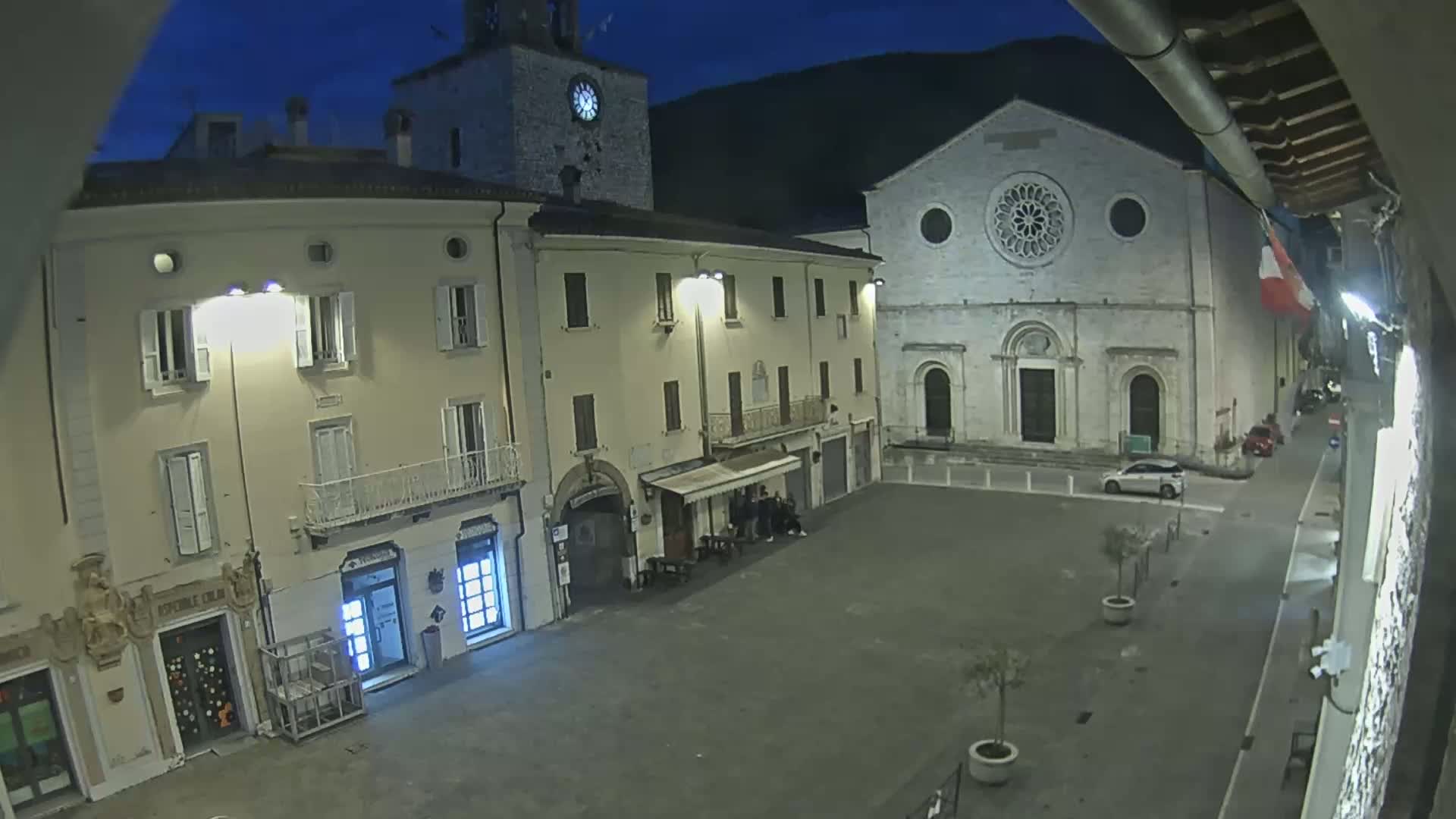 A nighttime view of a town square, featuring several buildings including a church with a large rose window, under a dark blue sky.