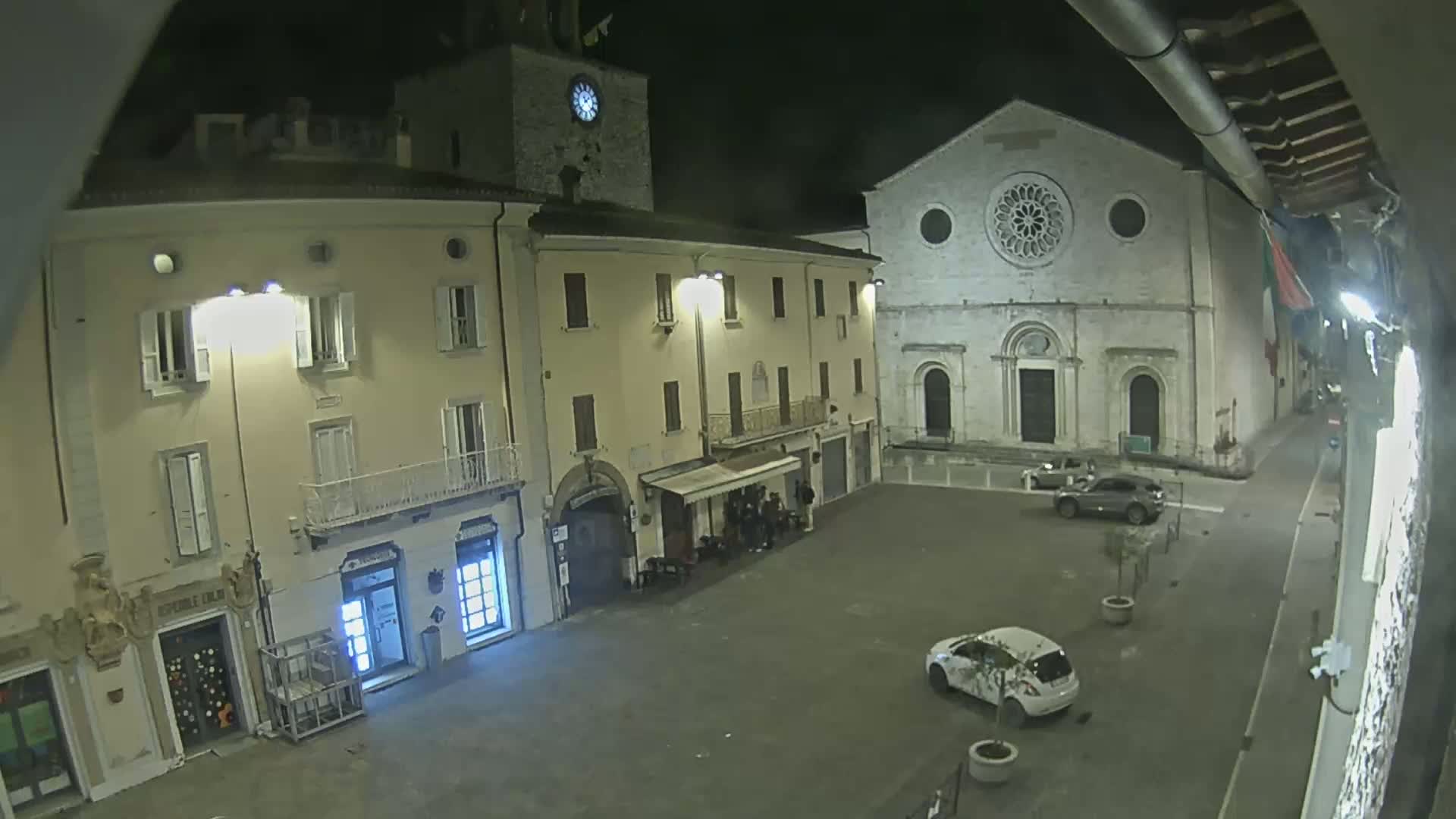 A nighttime view of a town square shows several buildings, including one with a clock tower and another with a large rose window, with a few cars parked in the empty space.