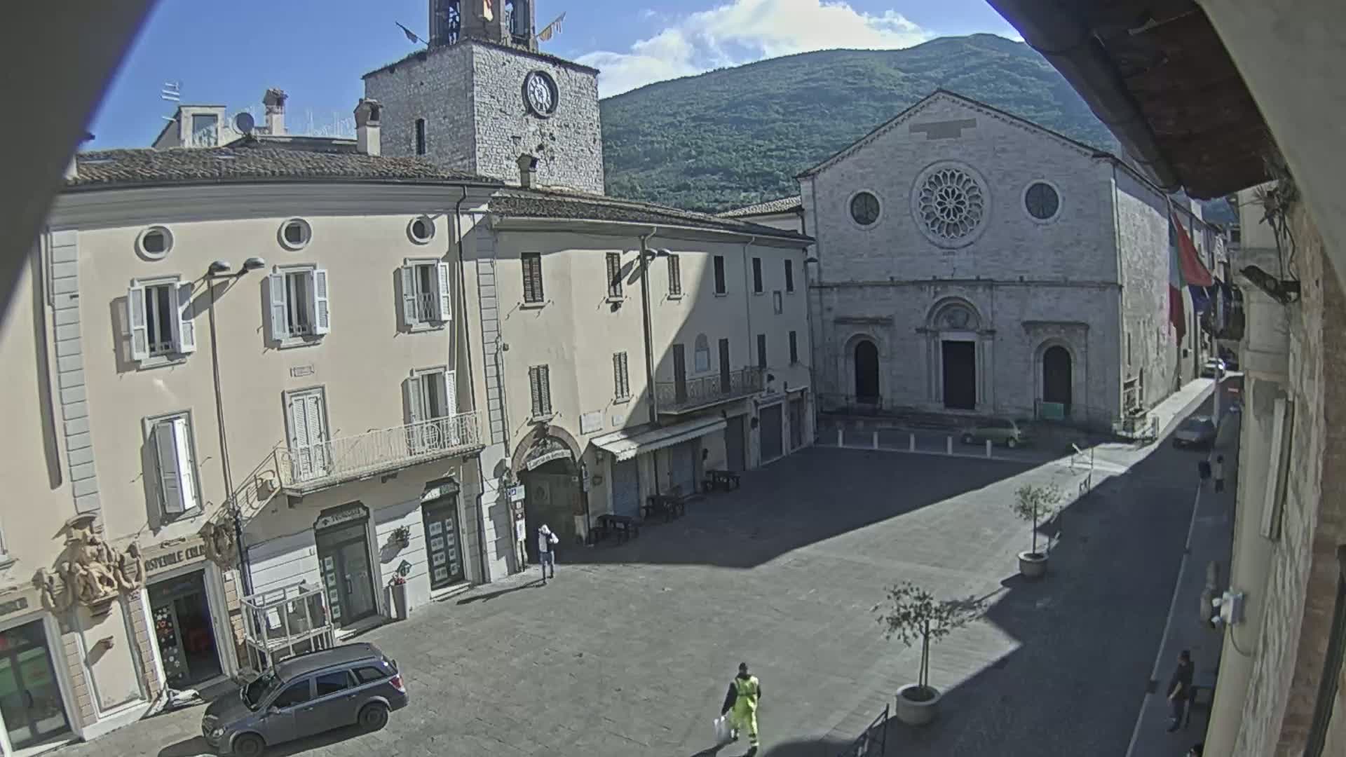 A sunny day reveals a town square with several stone buildings, including a clock tower and a church with a rose window, surrounded by sparsely populated streets.