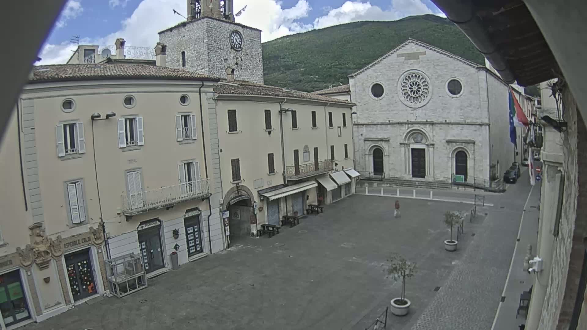 A mostly empty town square is surrounded by light-colored buildings, including a church with a rose window, under partly cloudy skies.