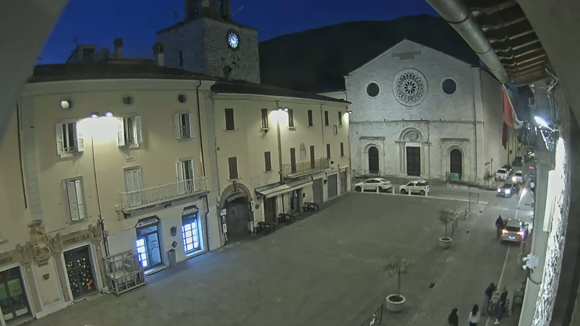 A nighttime view of an empty town square with several buildings, including a church with a large rose window, under a dark blue, clear sky.