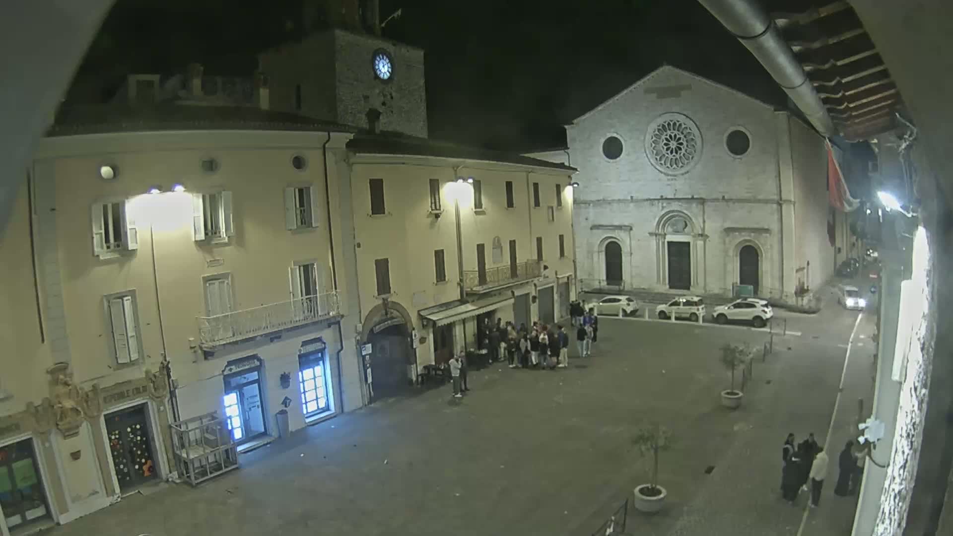 A nighttime view of a town square shows several people gathered near a building, with cars parked nearby and a large church in the background.