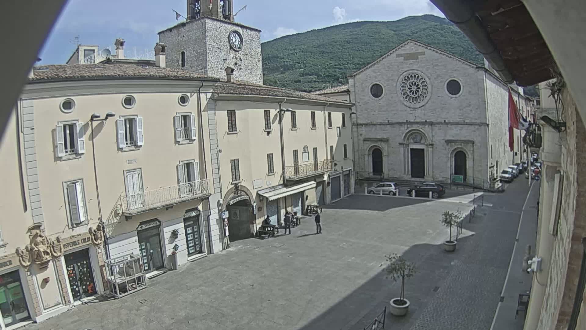 A sunny day reveals a town square with several buildings, including a church with a rose window, a clock tower, and multiple shops, with a few people scattered about.