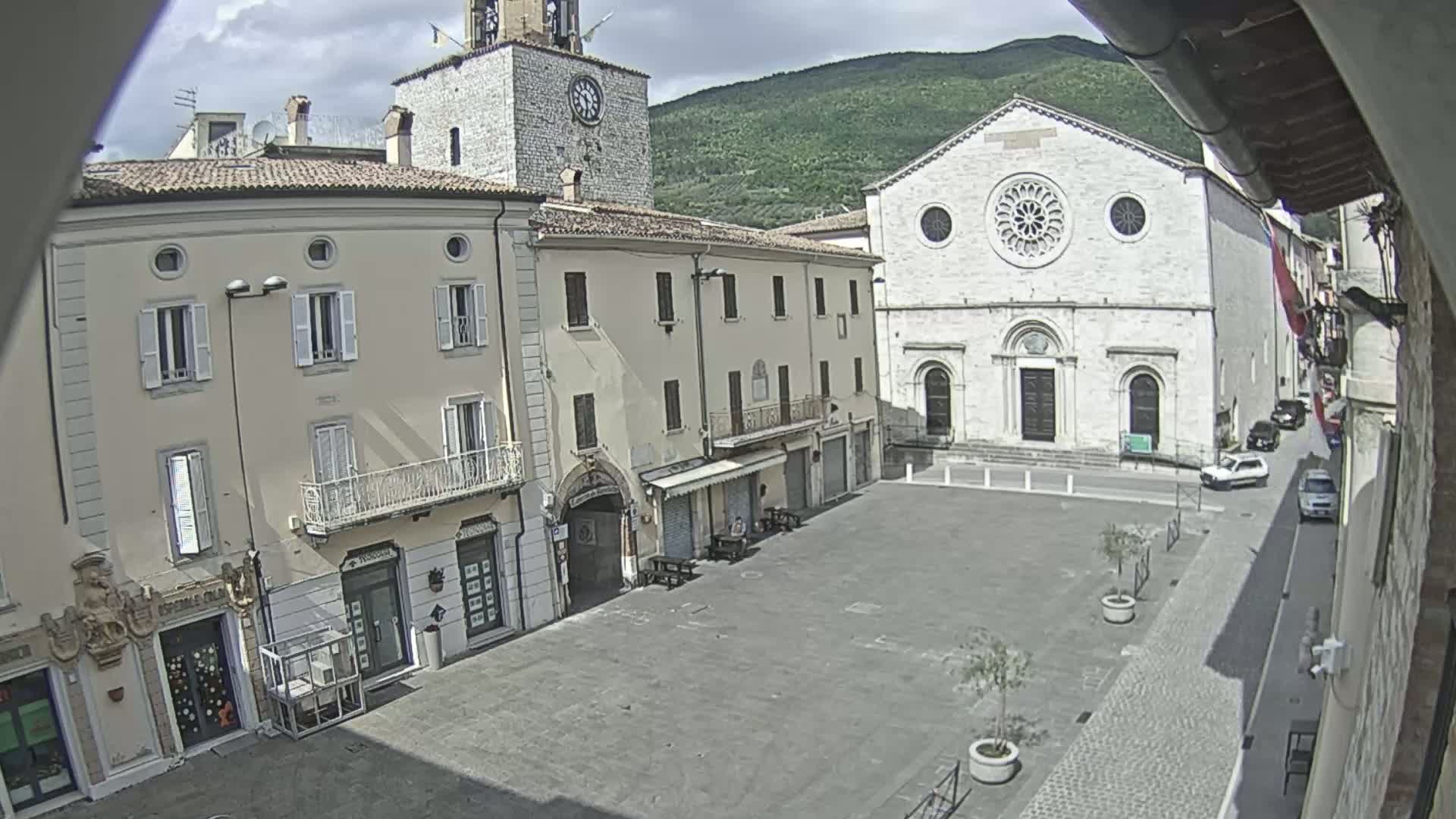 A mostly empty town square, under partly cloudy skies, is surrounded by buildings including a clock tower and a large stone church.