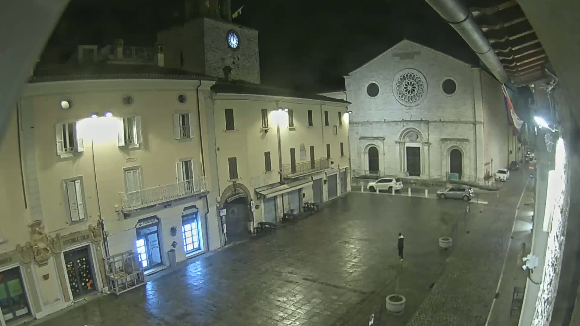 A wet, nighttime town square is shown, with buildings surrounding it including a clock tower and a large stone church with a rose window.