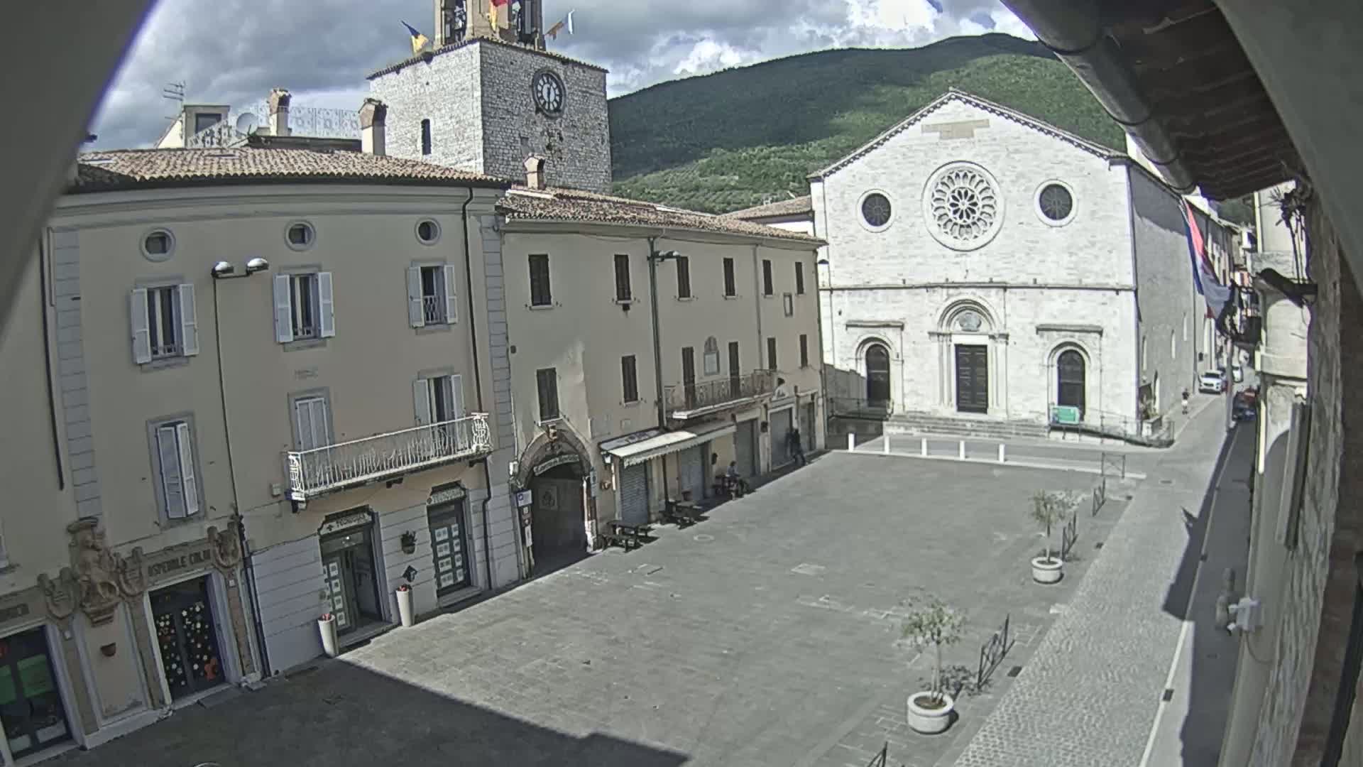 A mostly empty town square is situated between buildings, one of which is a church with a rose window, under a partly cloudy sky and mountains in the background.