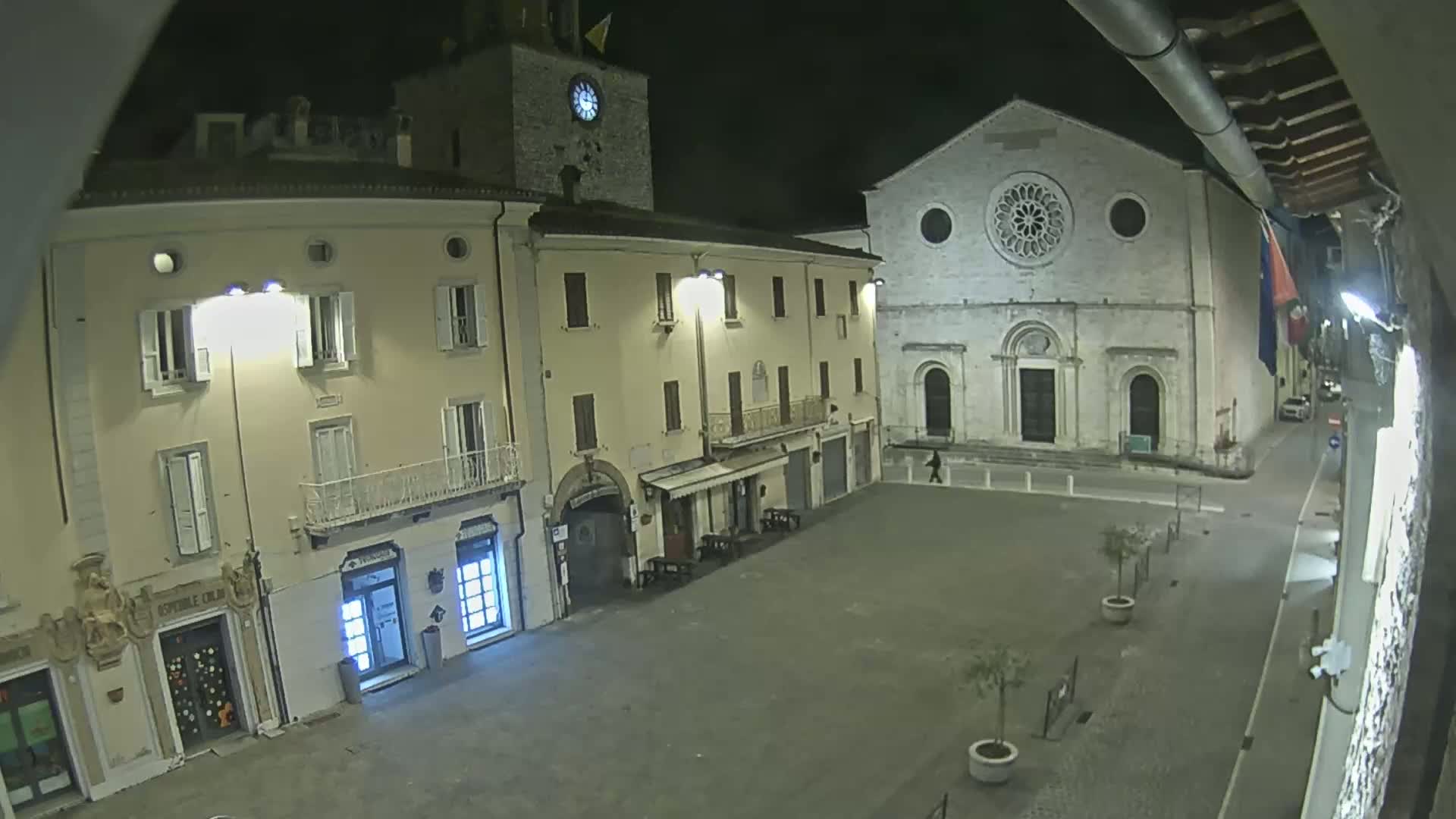 A nighttime view of a town square with buildings illuminated, including a clock tower and a church with a rose window.
