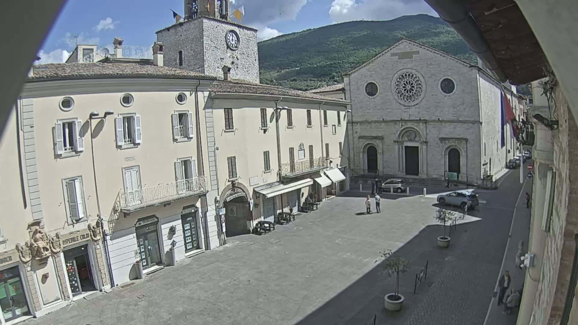 A sunny day reveals a town square, featuring several buildings, including a church with a rose window and a clock tower, along with a few people and parked cars.