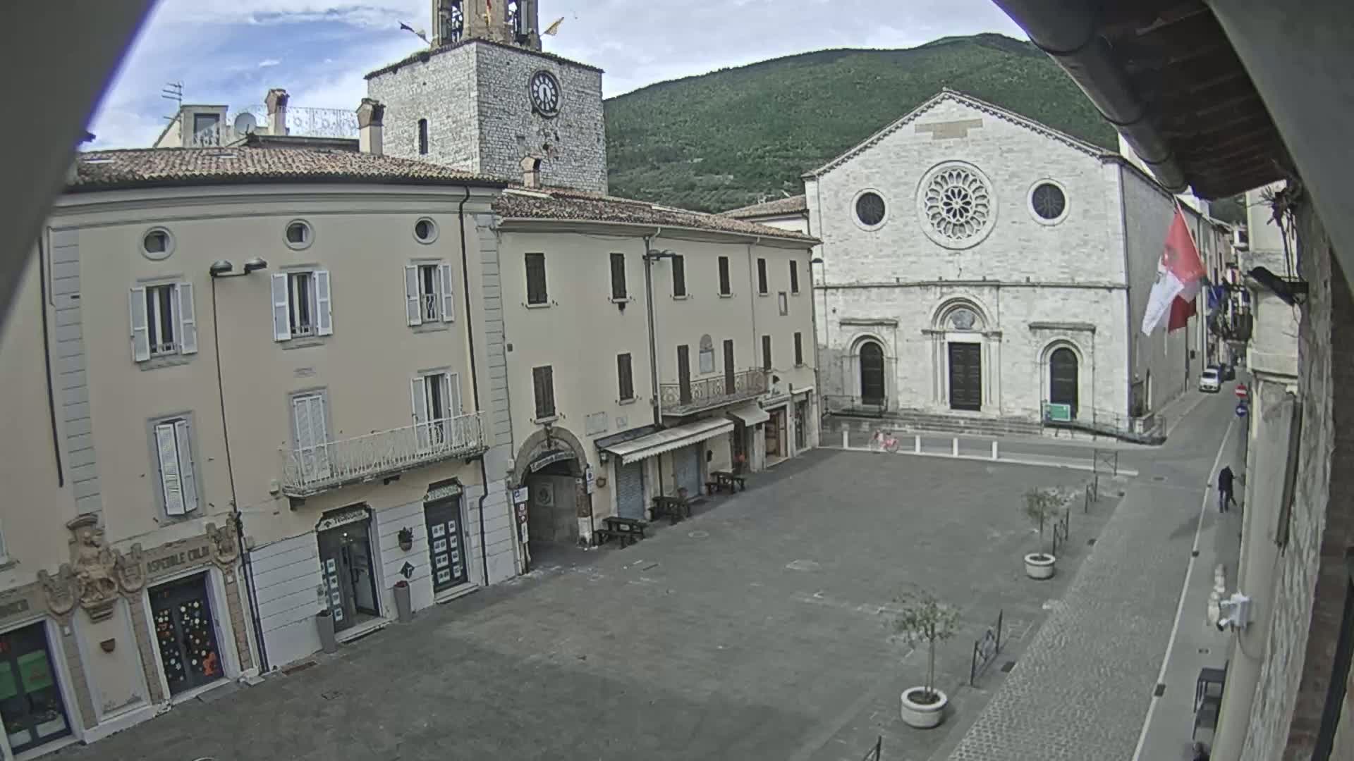 A mostly empty town square is surrounded by several light-colored buildings, including a church with a large rose window, under an overcast sky.