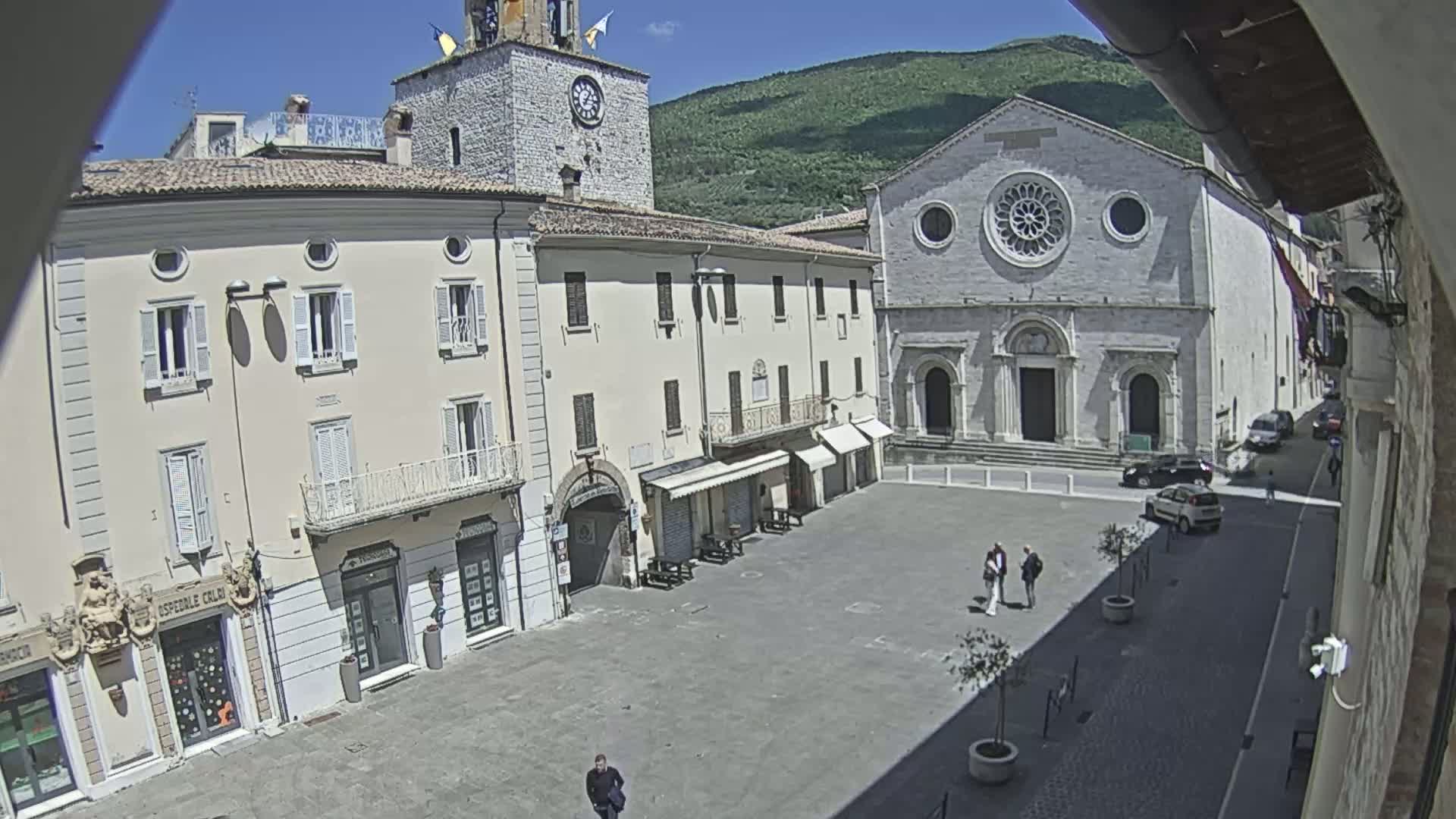 A sunlit town square, surrounded by light-colored buildings including a church with a rose window, is viewed from above on a clear day.