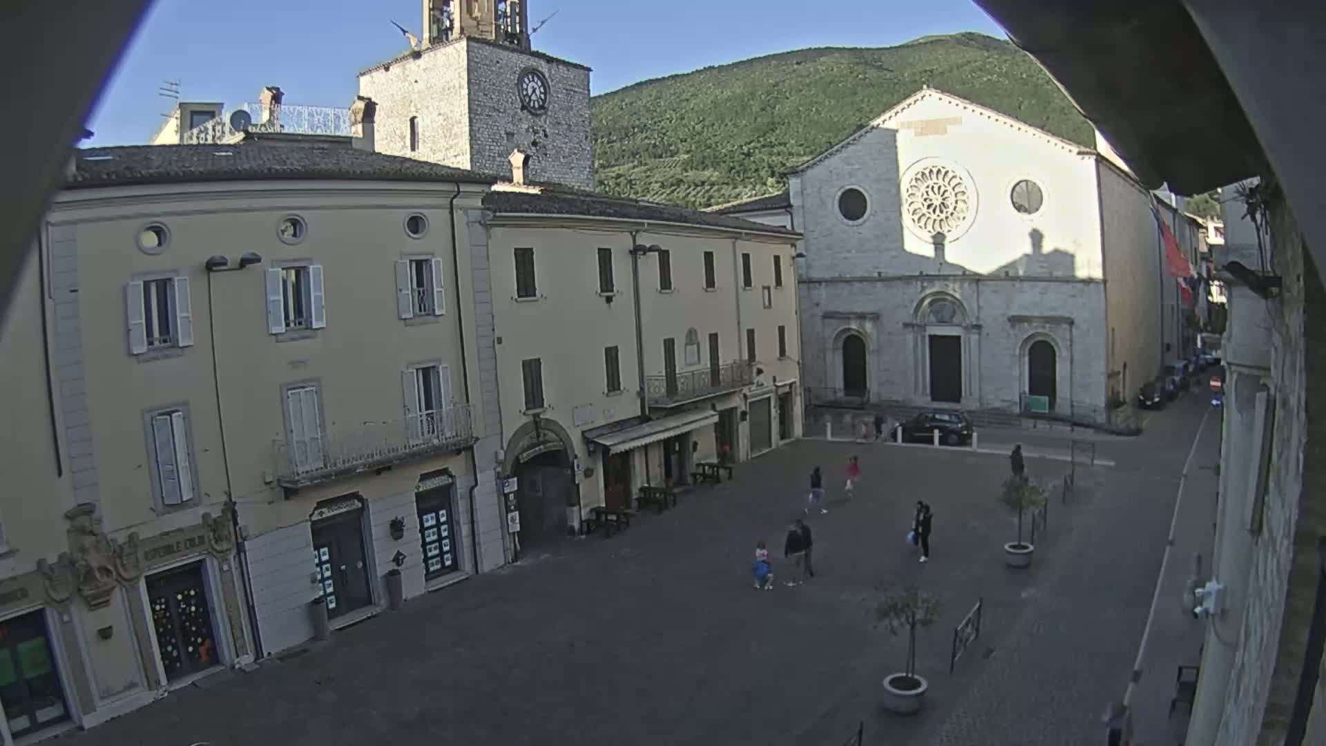 A mostly empty town square, under a clear sky, is surrounded by old stone buildings, including a church with a rose window and a clock tower.