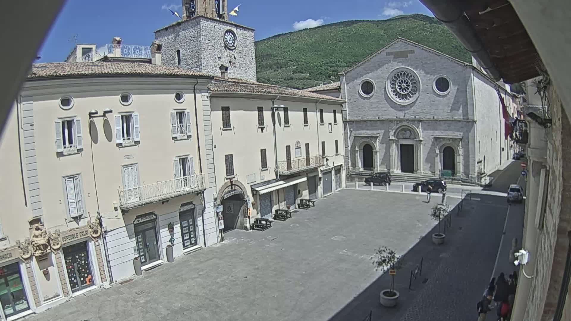 A sunlit town square is seen, featuring several multi-story buildings, including a church with a large rose window, under a mostly clear sky with a mountain in the background.