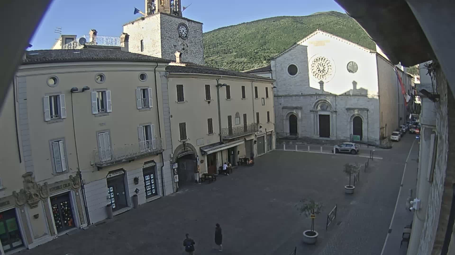 A mostly empty town square is viewed from above on a sunny day, with several buildings surrounding it, including a church with a rose window and a clock tower.