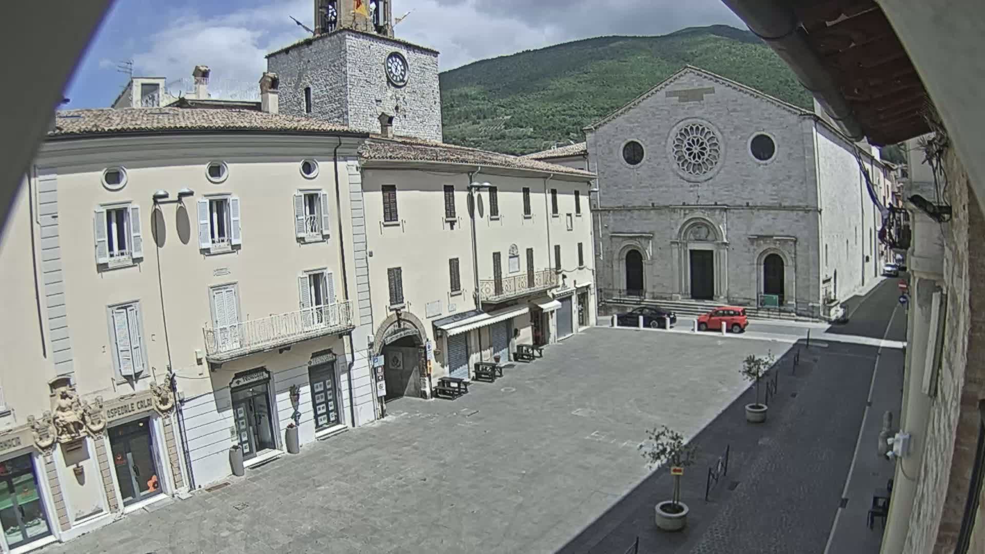 A sunny town square is seen from above, with several buildings including a clock tower and a church, under partly cloudy skies and mountains in the background.