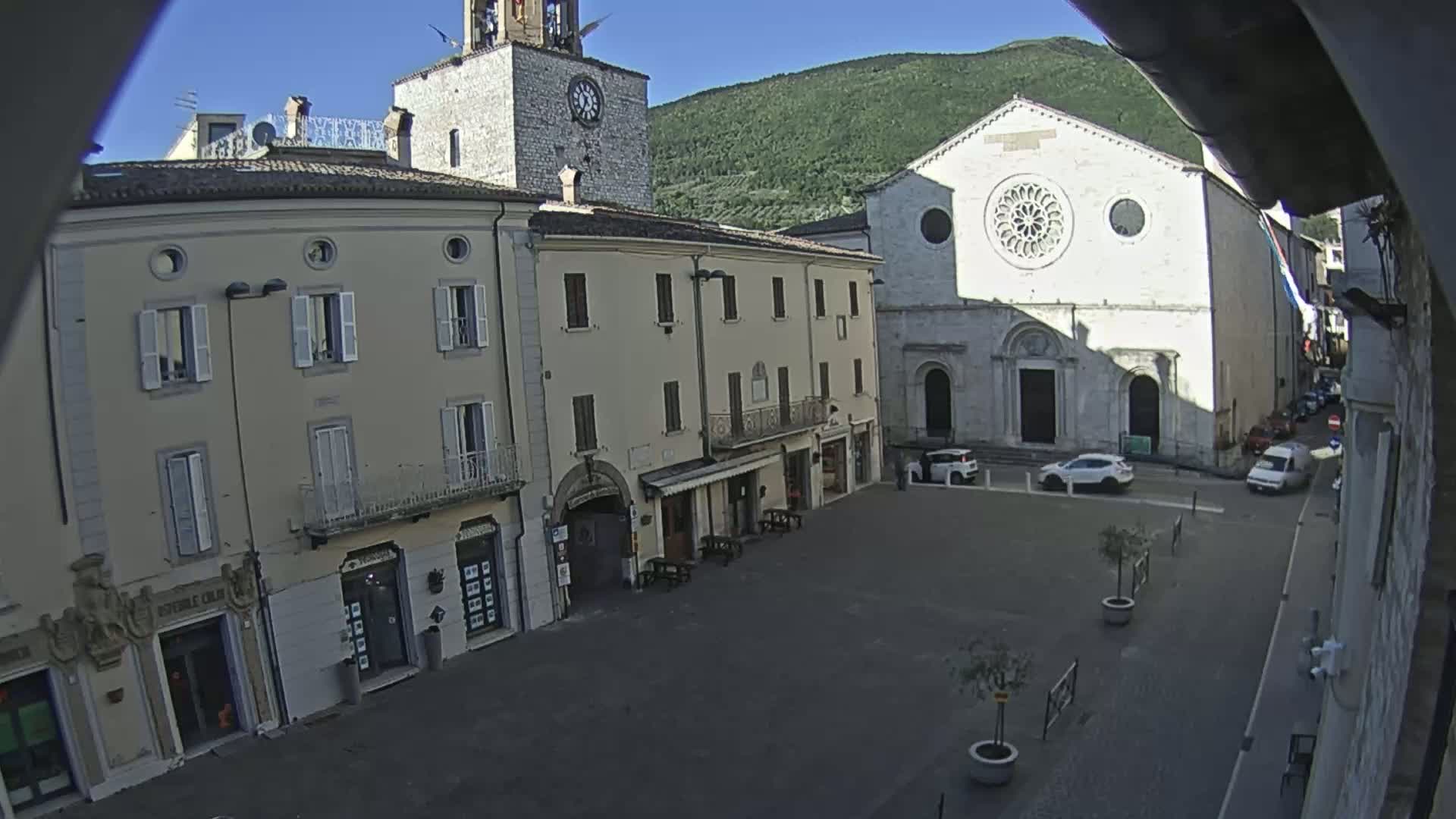 A sunny town square with several light-beige buildings, including a church with a large rose window, and a clock tower.