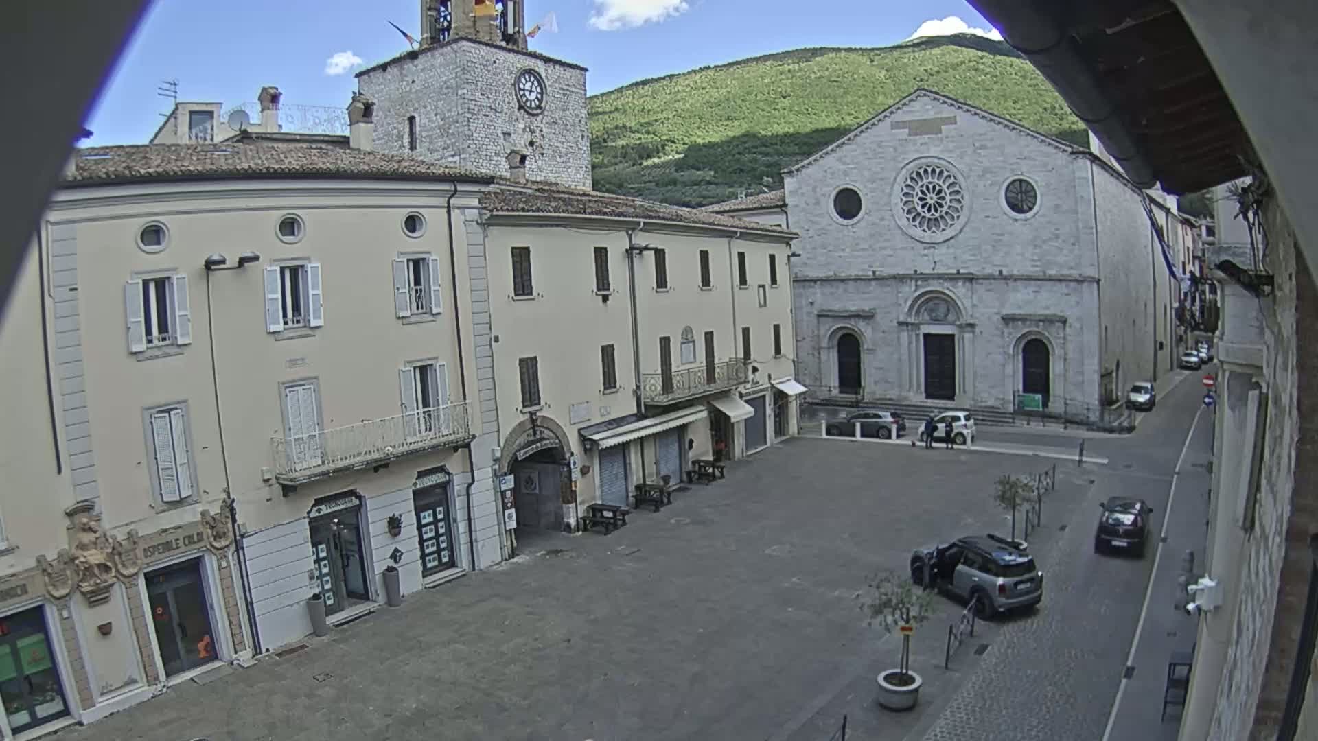 A mostly empty town square is seen on a sunny day, with several buildings, including a church with a rose window, and a few cars parked nearby.