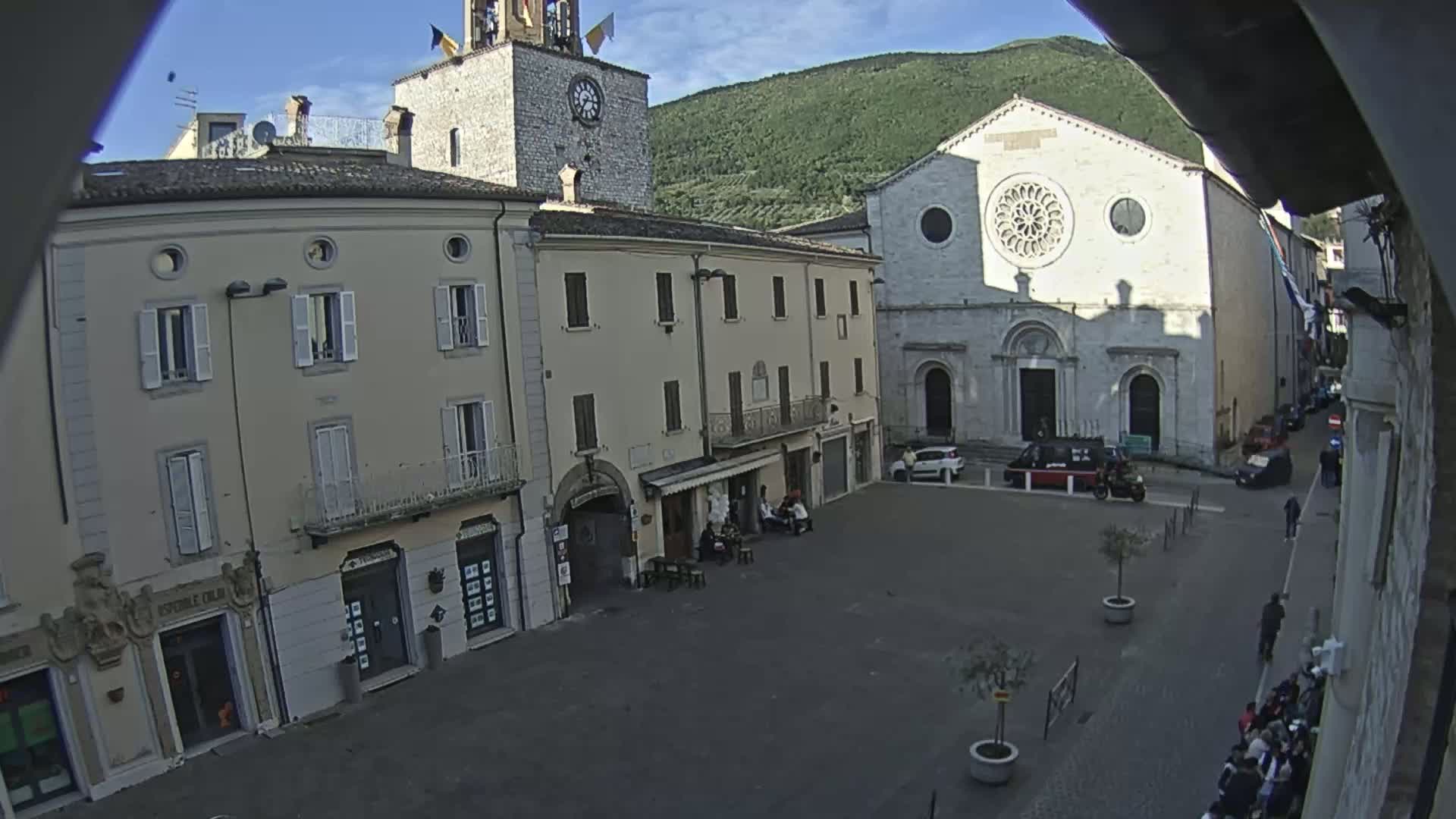 A town square with several multi-story buildings, including a church with a rose window, under a mostly sunny sky.