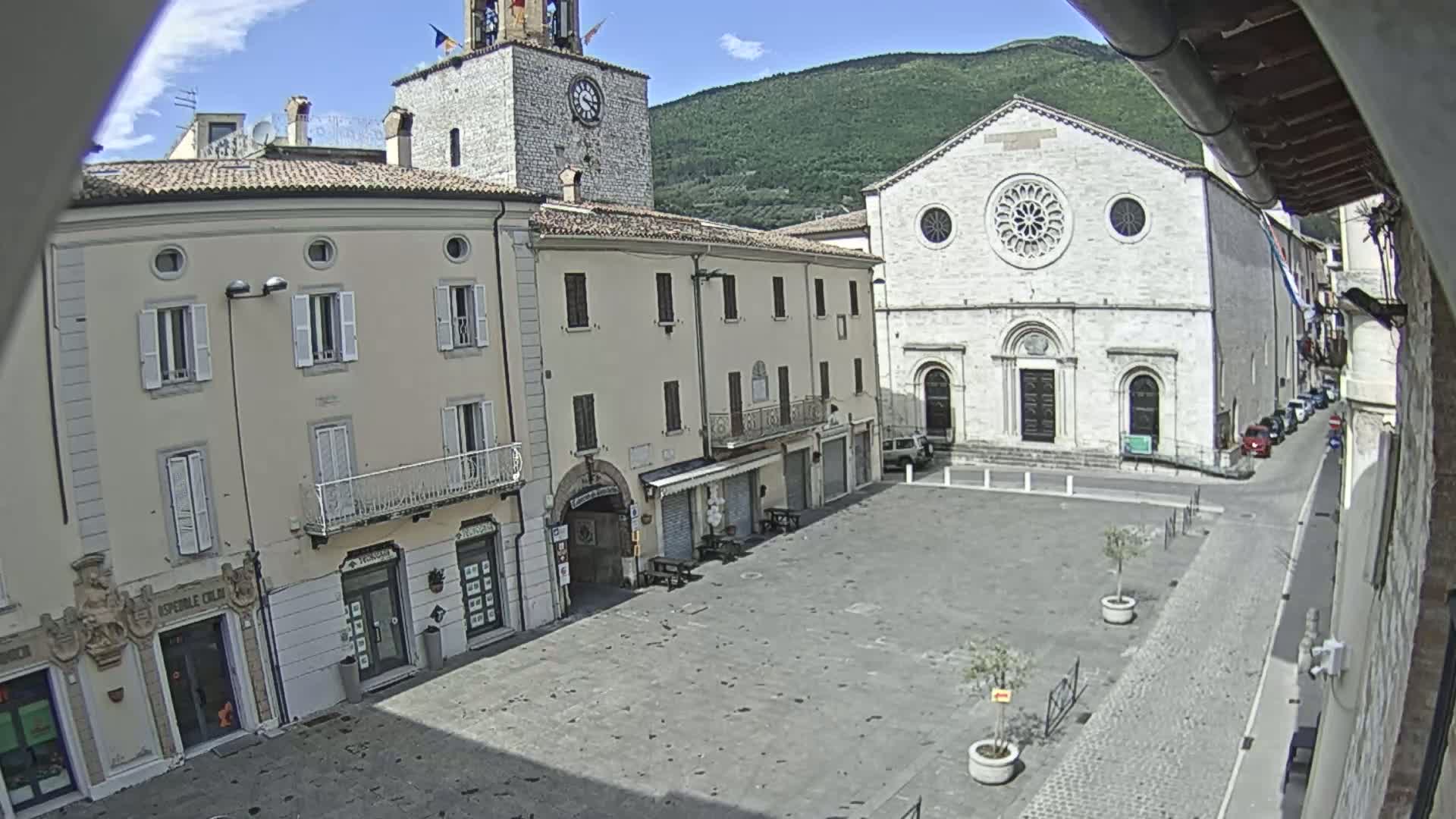 A sunny day in a town square, featuring several light-colored buildings, including a church with a rose window, a clock tower, and shops, with a few cars parked nearby.