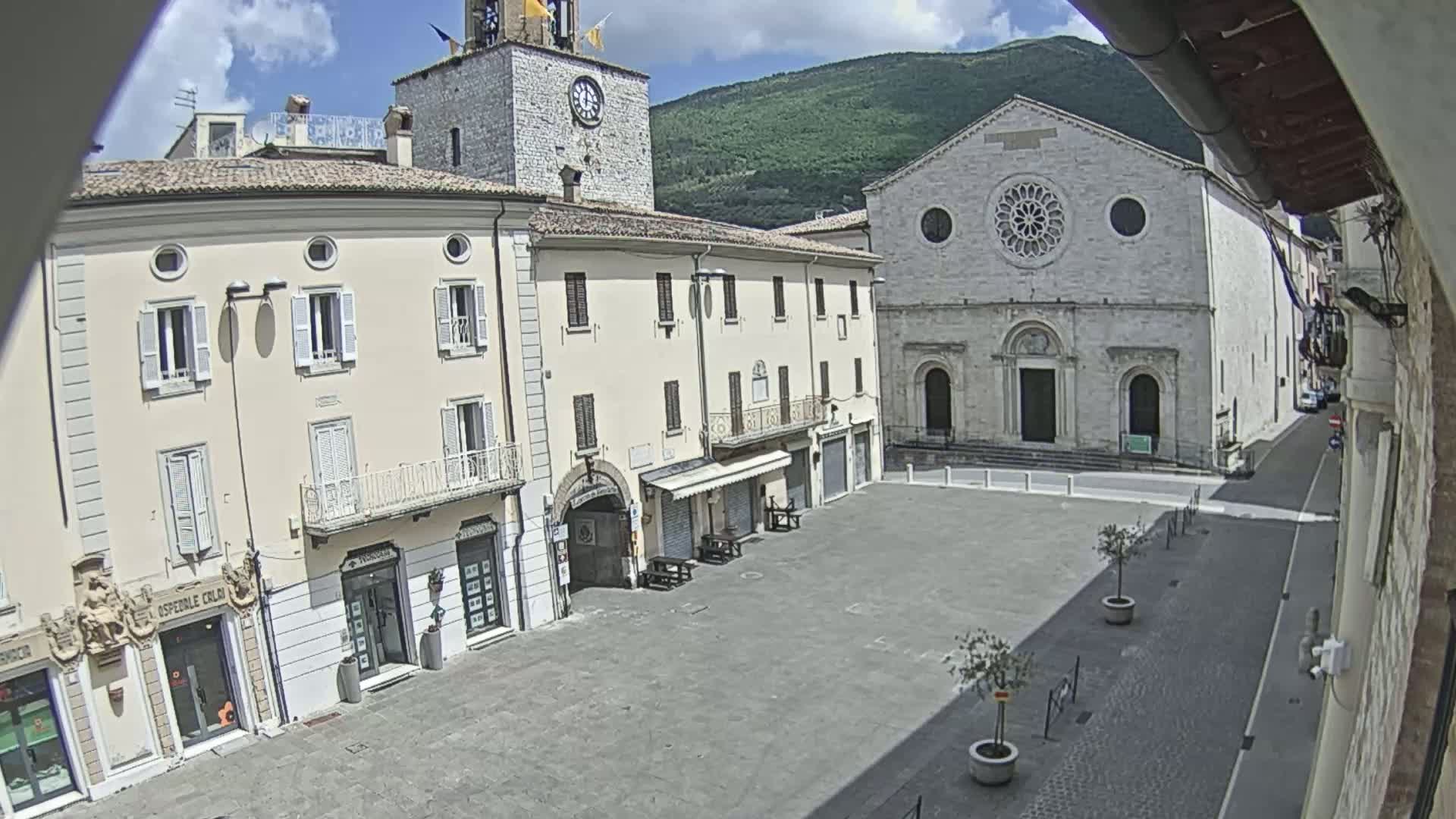 A mostly empty town square on a sunny day, surrounded by light-colored buildings including a church with a rose window and a clock tower.