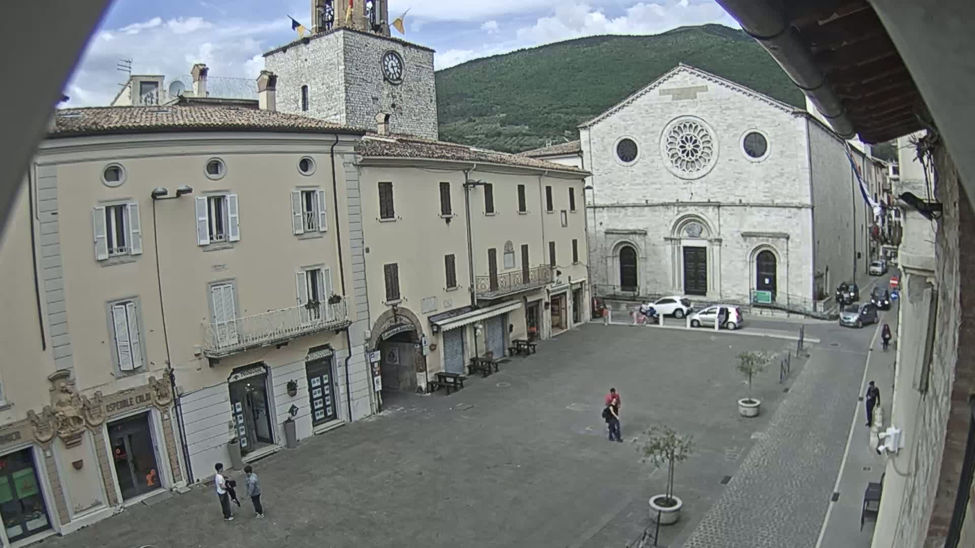 A mostly empty town square is seen on a partly cloudy day, with several buildings surrounding it, including a church with a rose window and a clock tower.