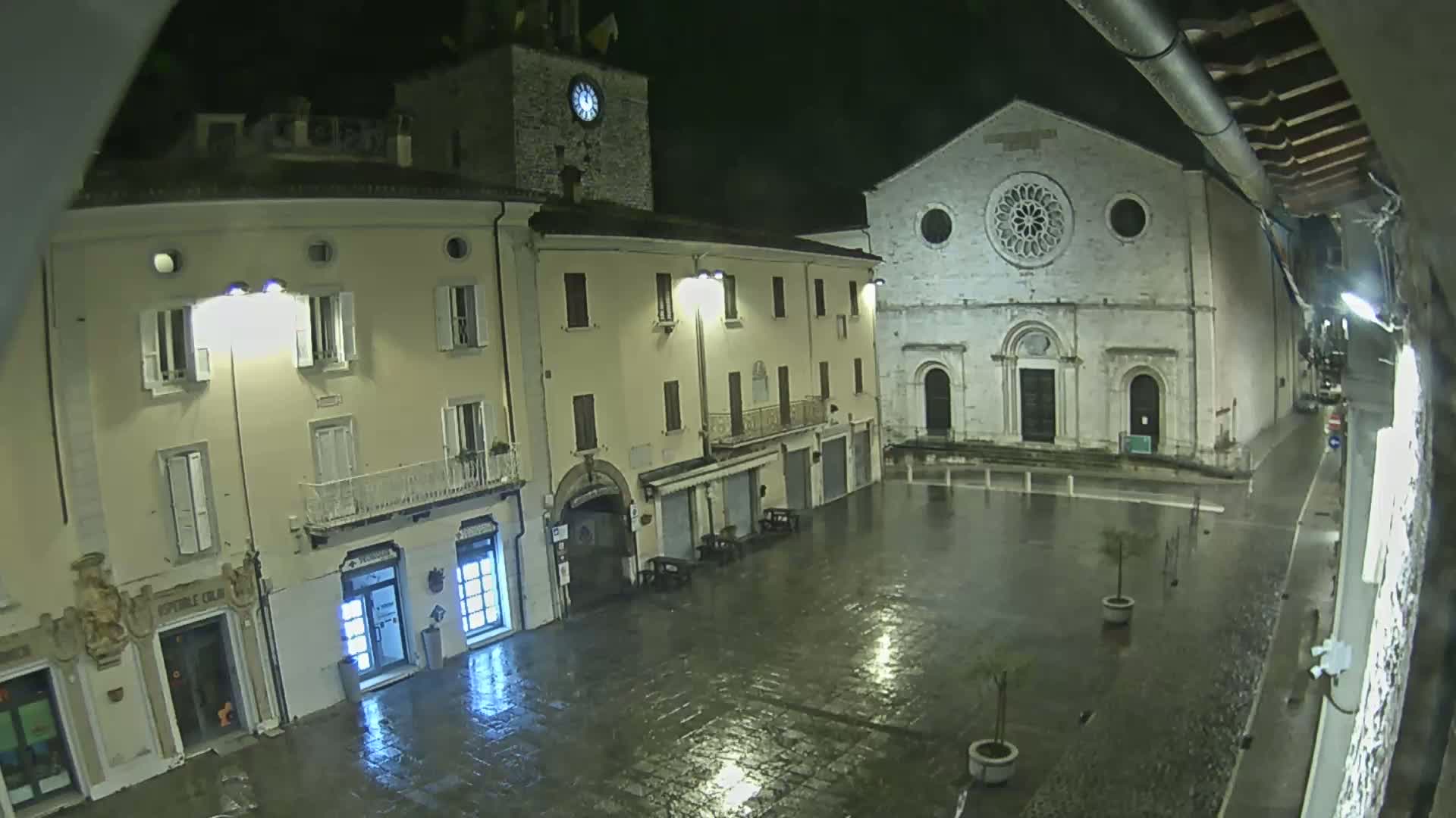 A rain-slicked town square at night is illuminated by streetlights, showing several buildings including a large church with a rose window and a clock tower.