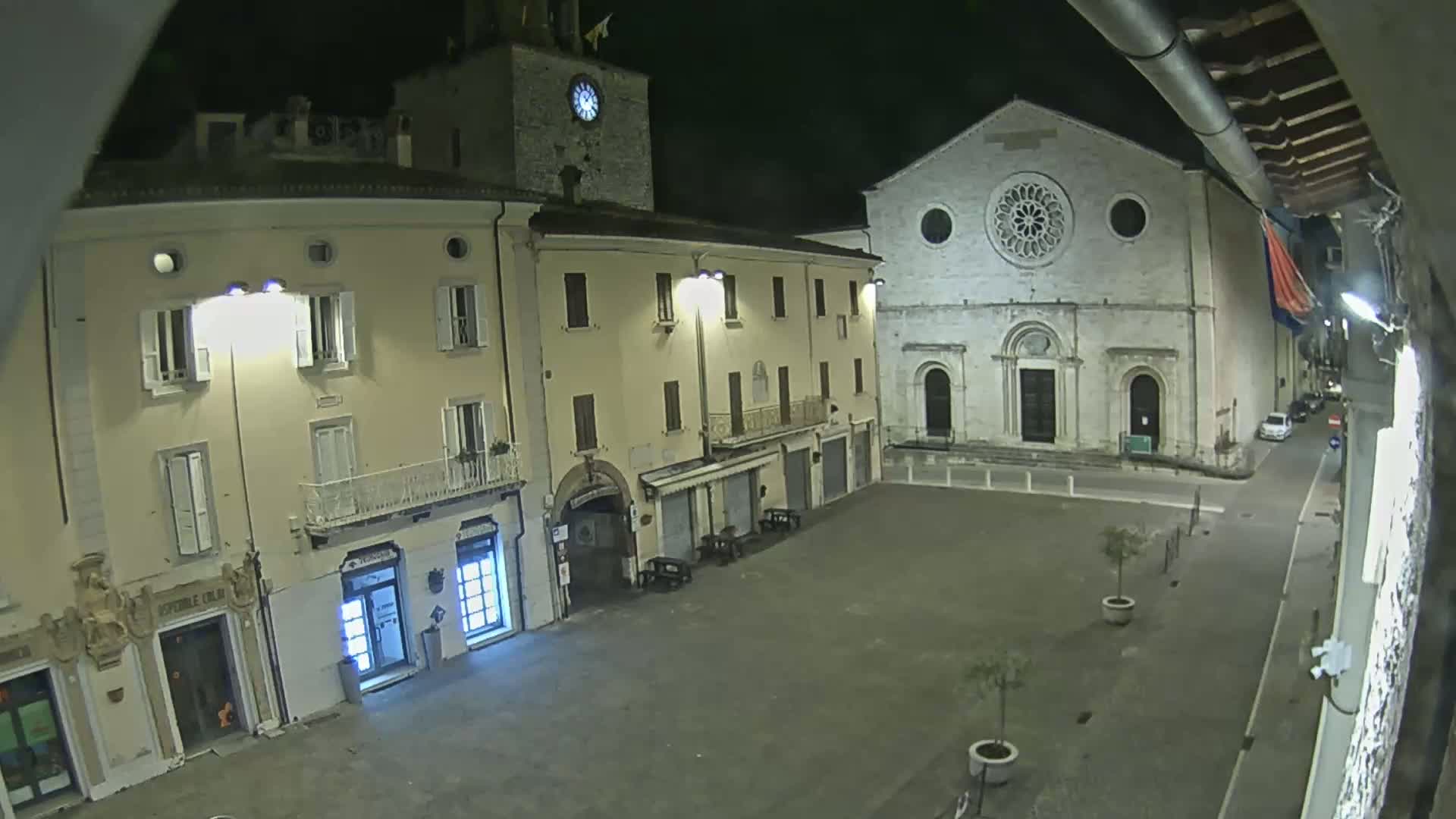 An empty, stone town square at night, illuminated by streetlights, features buildings including a church with a rose window and a clock tower.