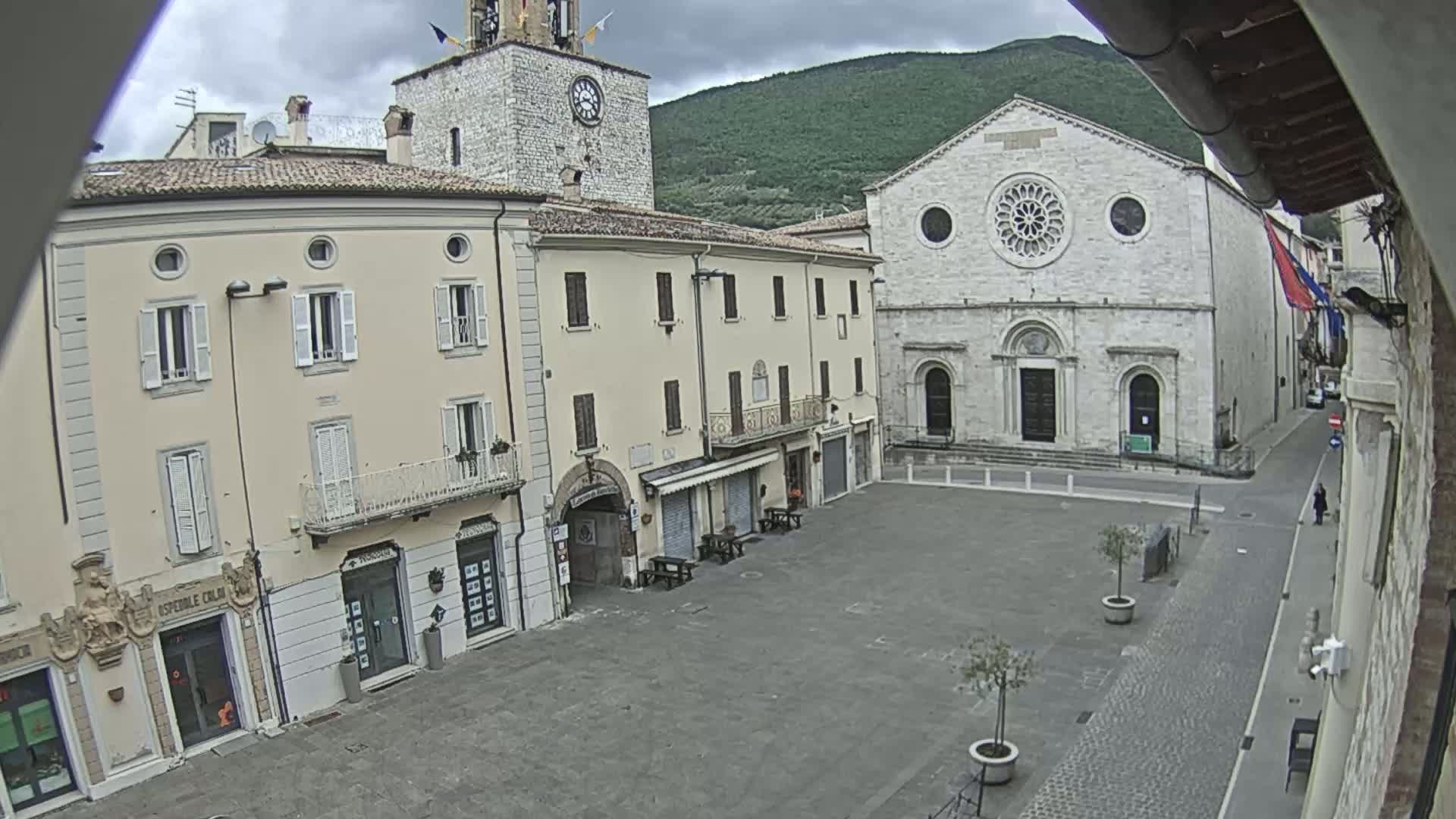 A mostly empty town square is viewed from above on an overcast day, showing several light-colored buildings including a church with a large rose window and a clock tower.