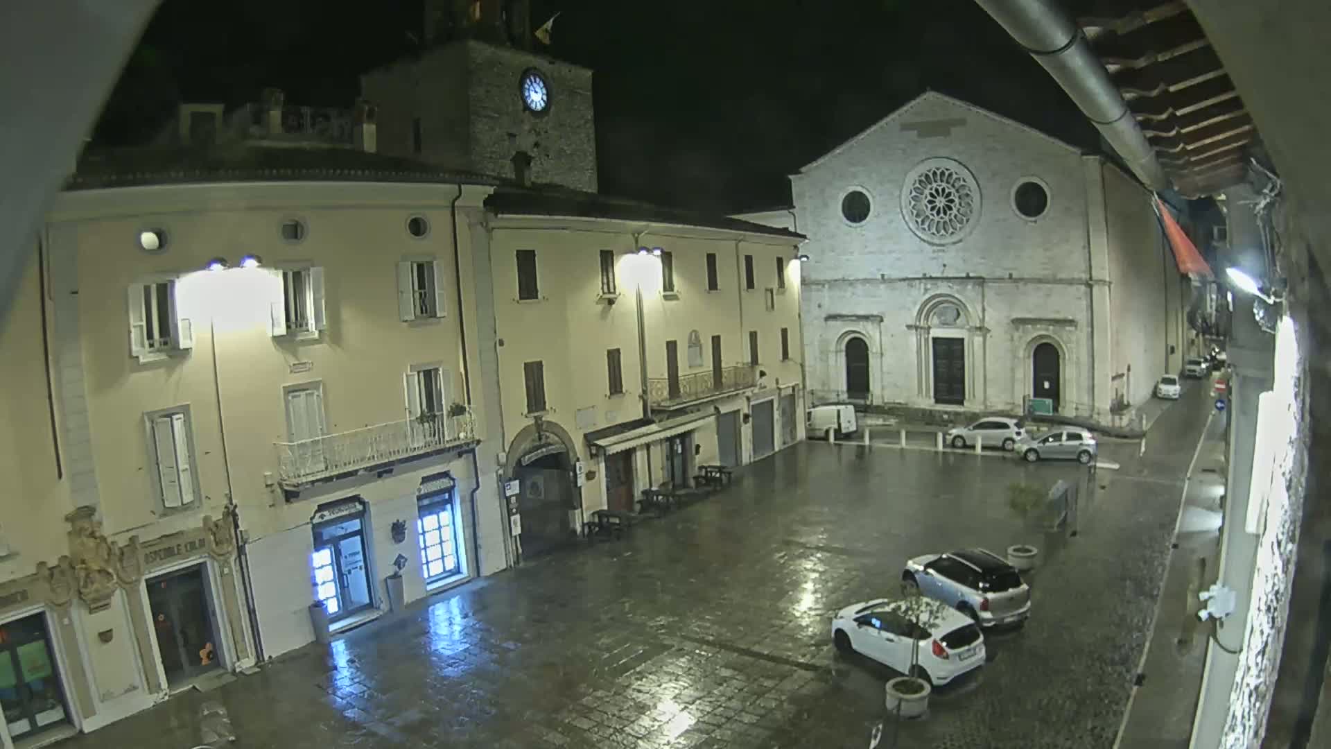A rain-slicked town square at night, featuring several buildings including a church with a large rose window, and a few parked cars.