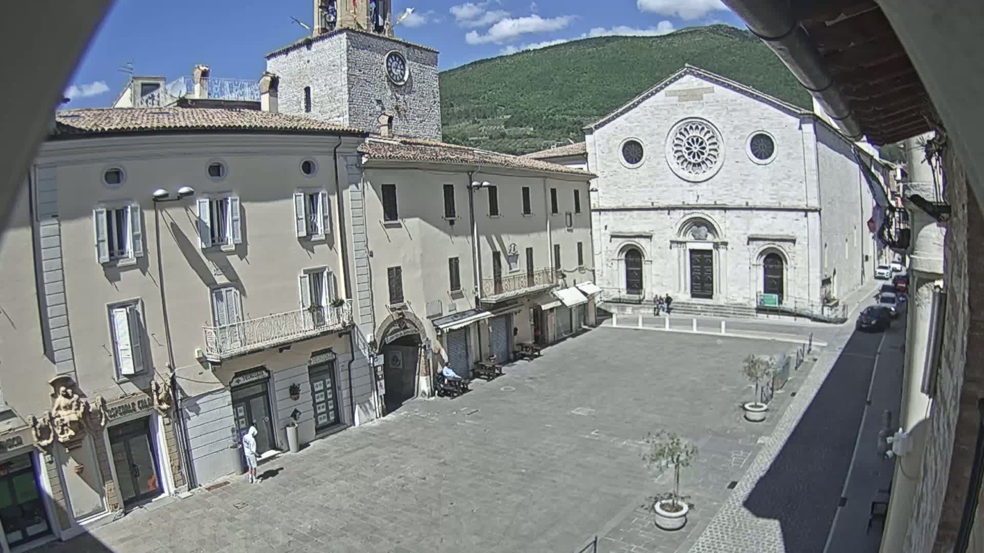 A sunny town square is surrounded by light-colored buildings, including a church with a large rose window and a clock tower.
