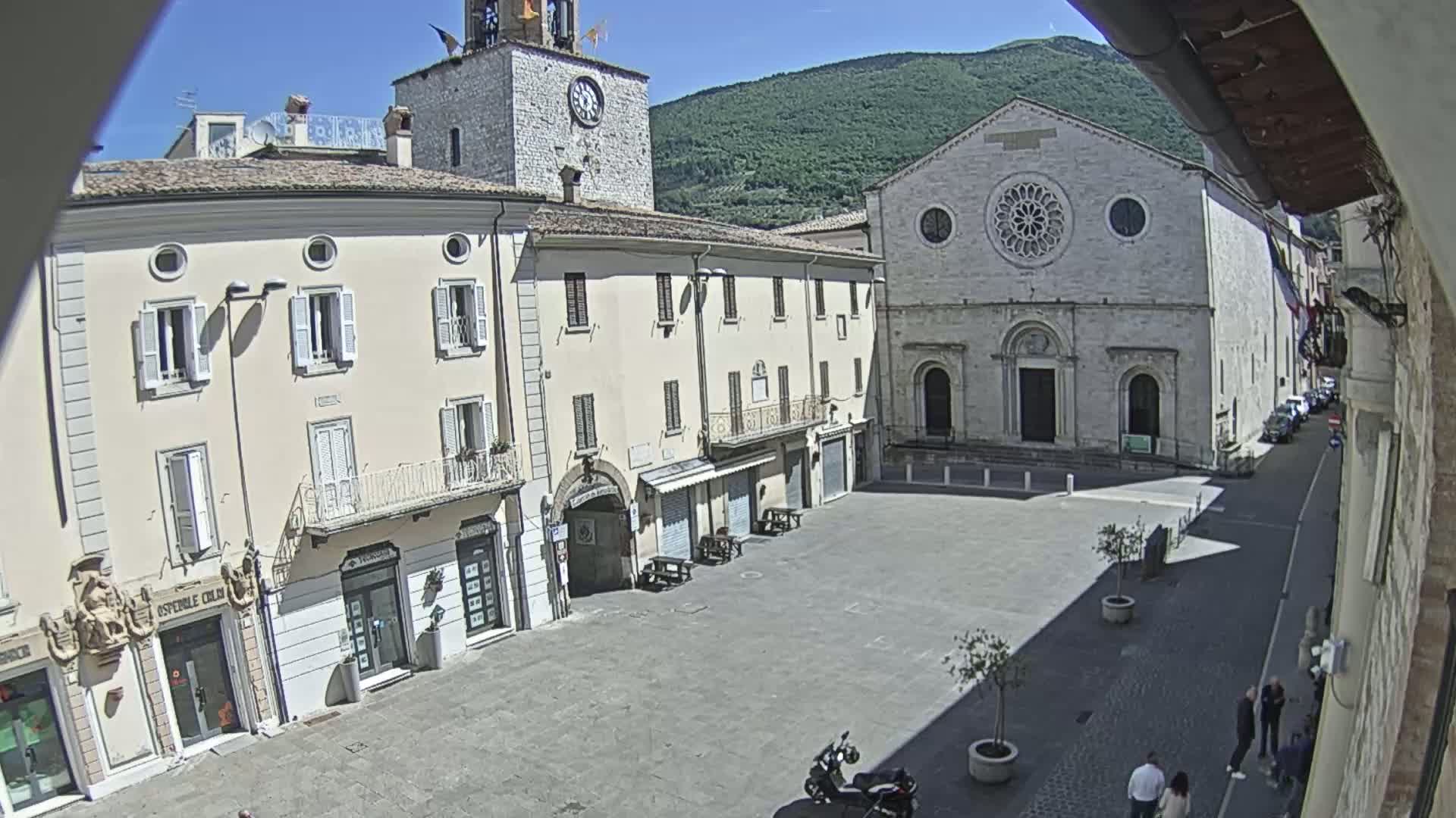 A sunny town square with light-colored buildings, including a church with a large rose window, is seen from an elevated vantage point.