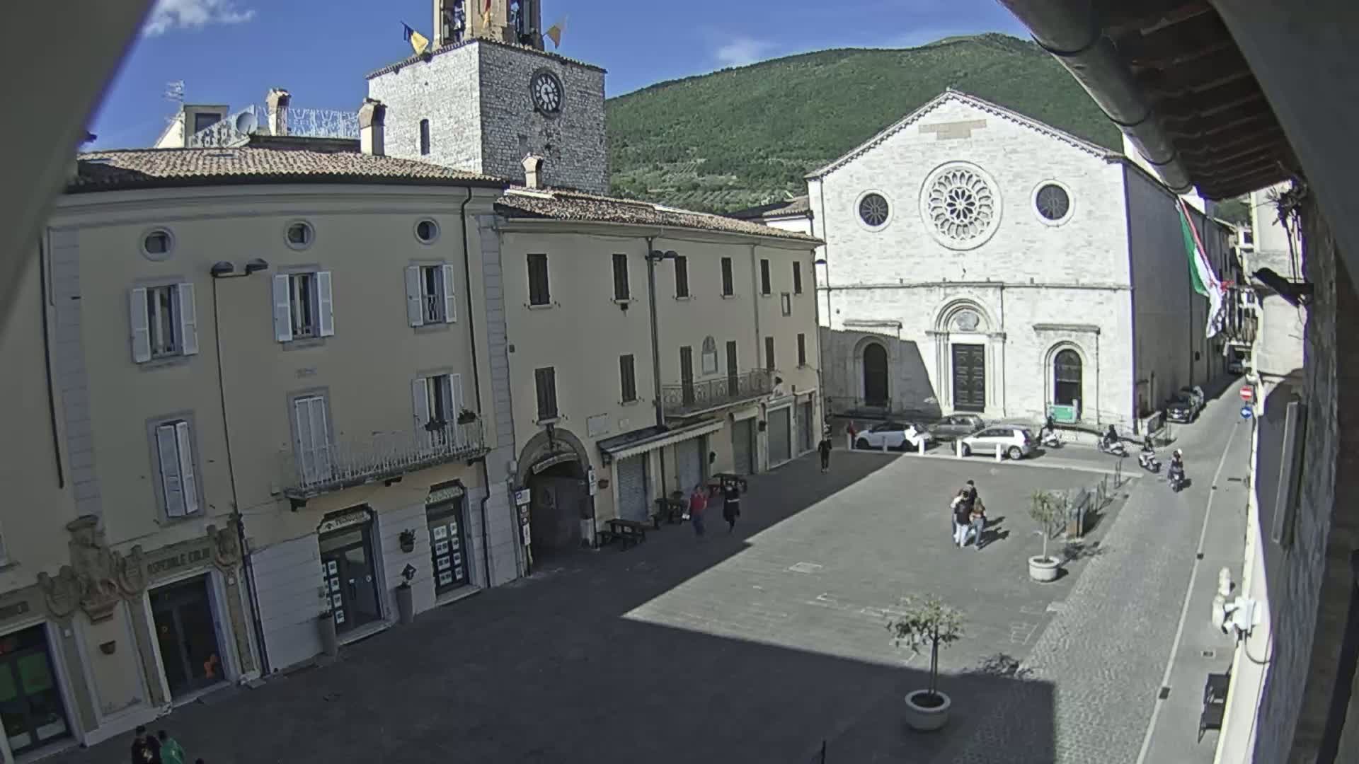 A sunny day in a town square shows several buildings, including a church with a large rose window, and a few pedestrians and parked cars.