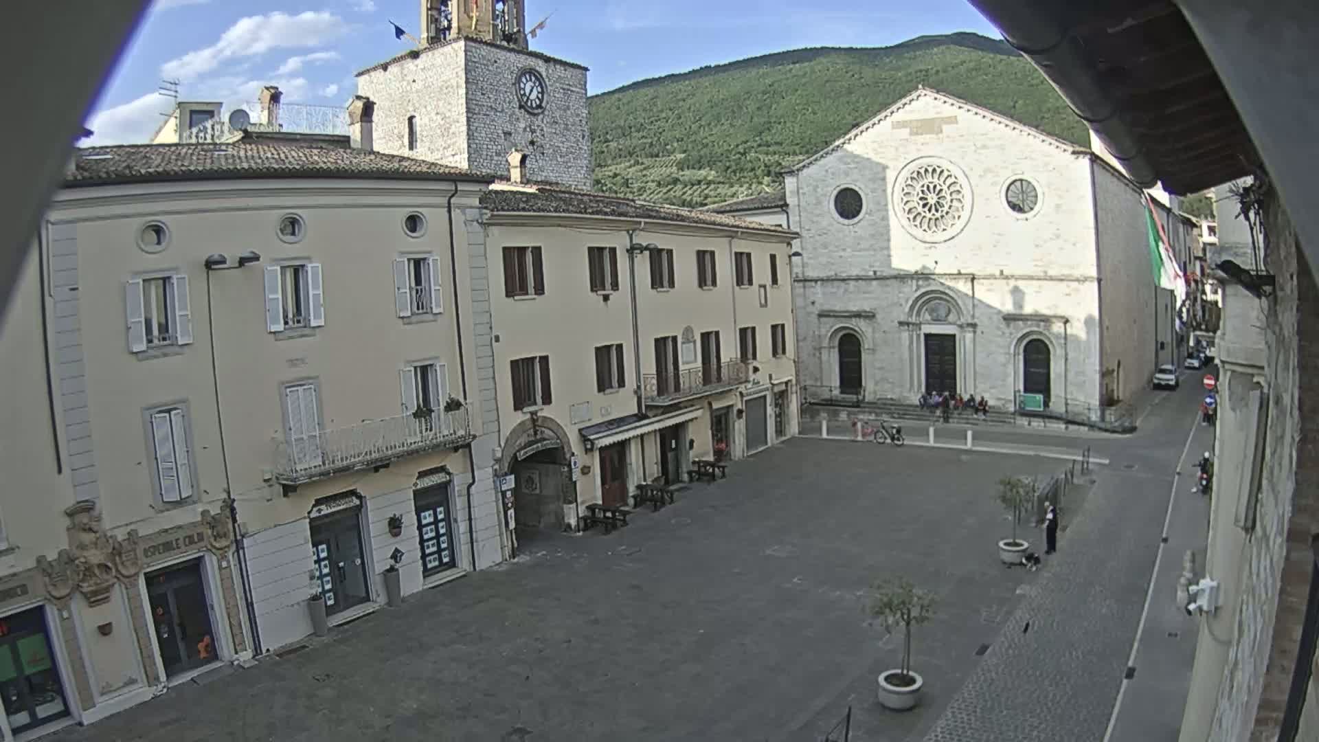 A town square with several light-colored buildings, including a church with a large rose window, under a partly cloudy sky and mountains in the background.
