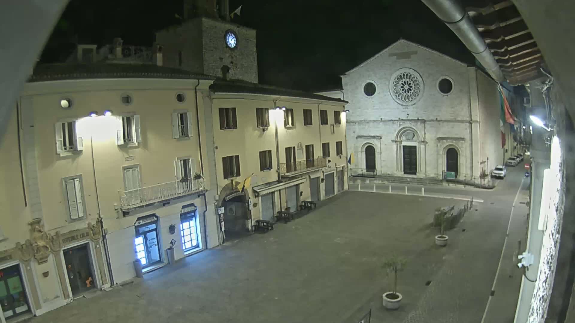 A nighttime view of a town square shows several buildings, including a church with a rose window, and a clock tower, under clear skies.