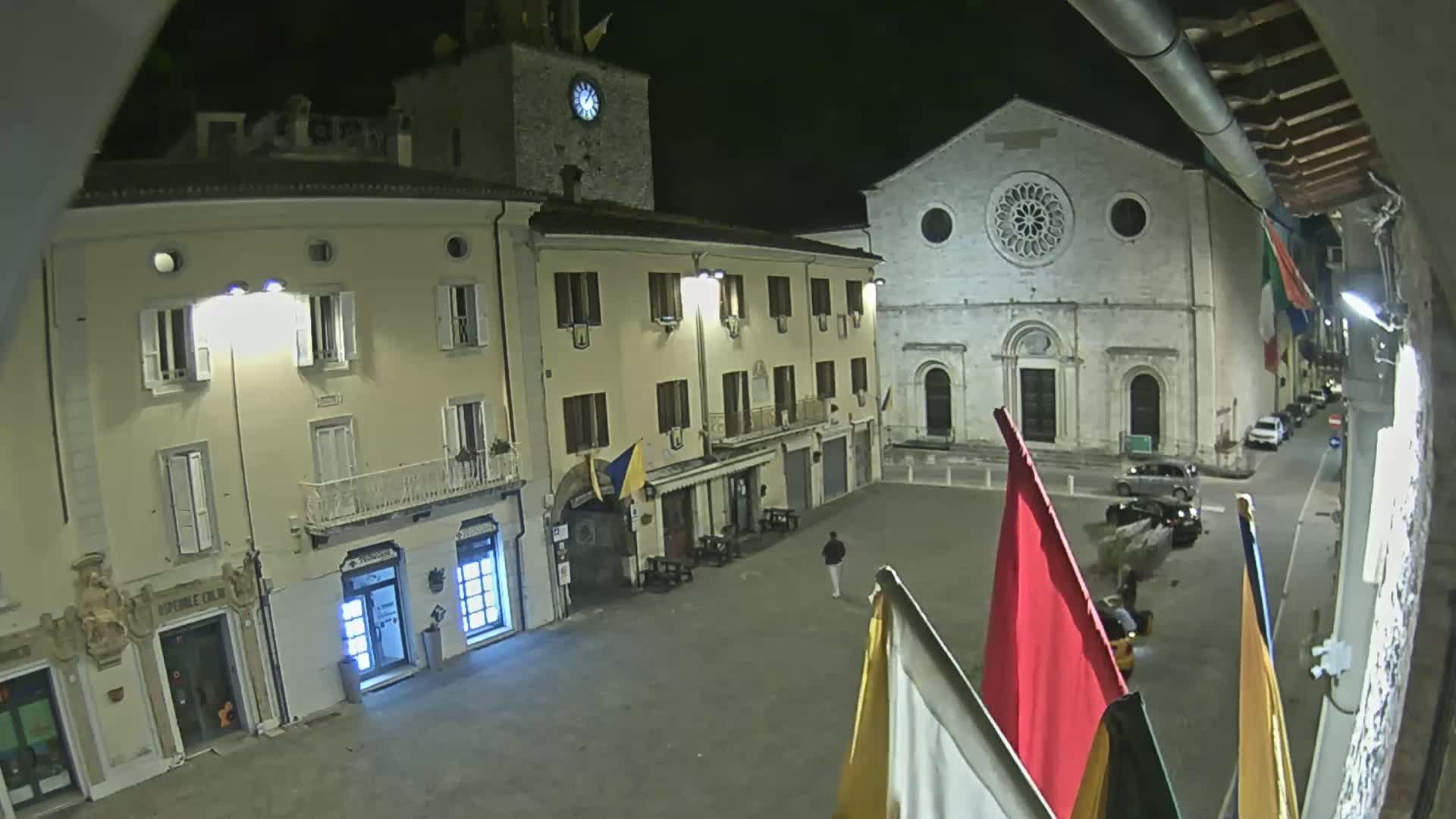 A nighttime view of a town square with several buildings, including a church with a rose window, and a person walking across the square.
