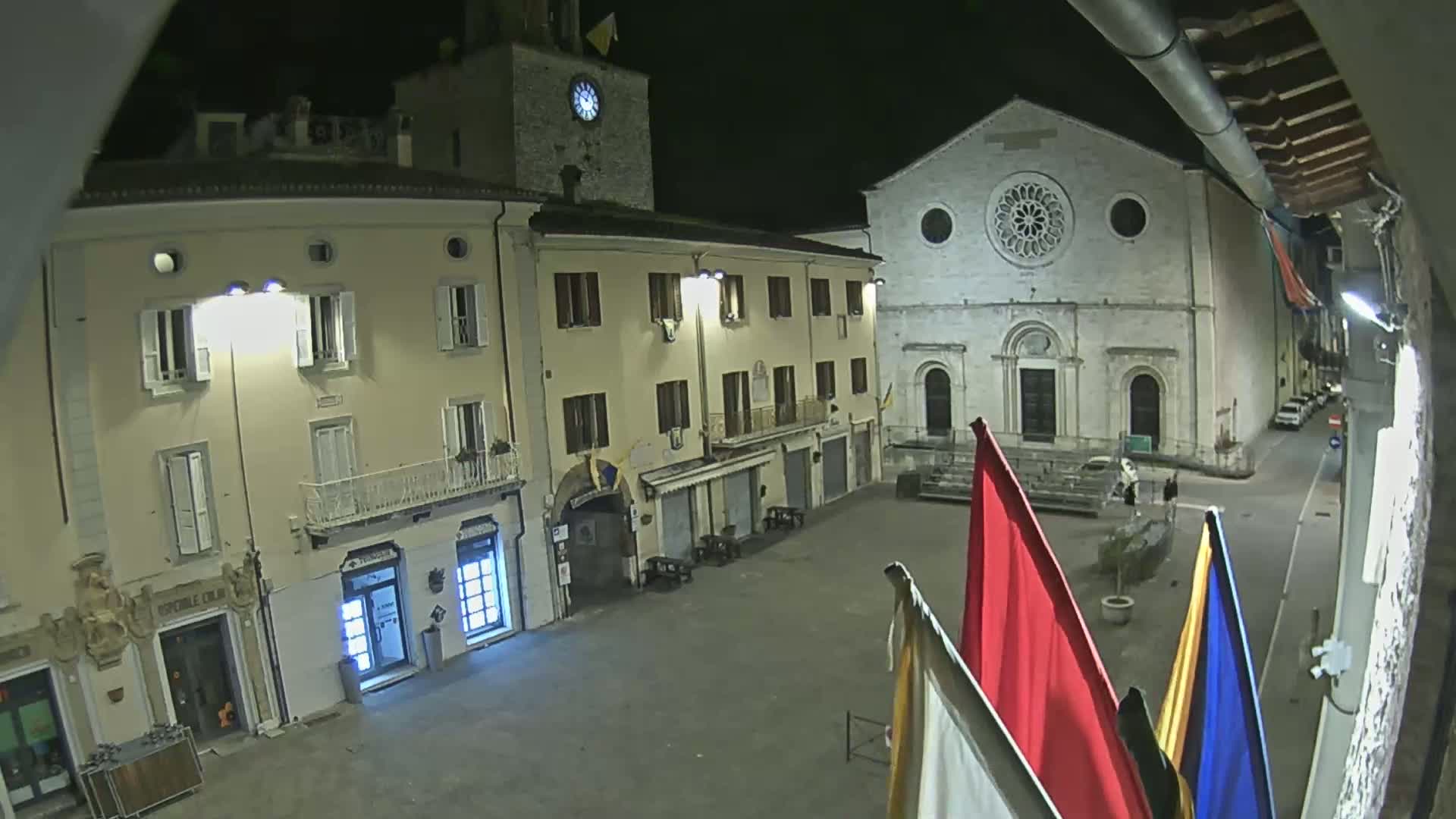 A nighttime view of a town square with buildings lit by artificial light, including a church with a rose window, and a few flags in the foreground.