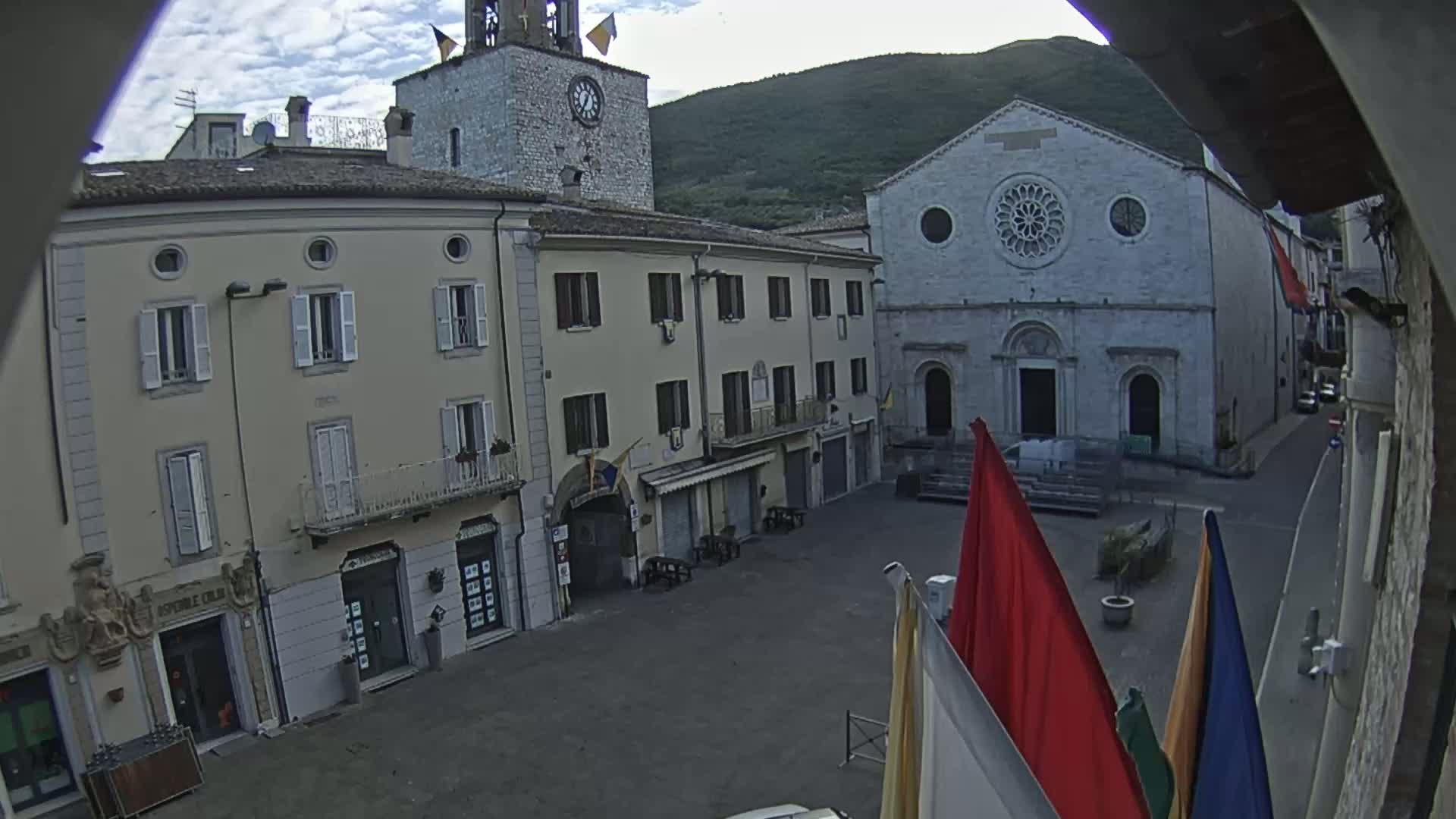 A town square is seen on an overcast day, with stone buildings, including a church with a rose window, surrounding it; several flags stand in the square.