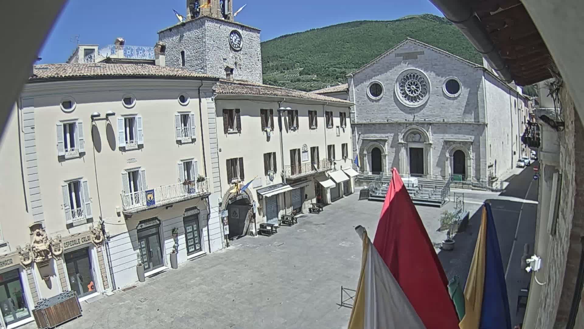 A sunny day reveals a town square with light-colored buildings, including a church with a rose window, and a clock tower, all nestled against a backdrop of green hills.