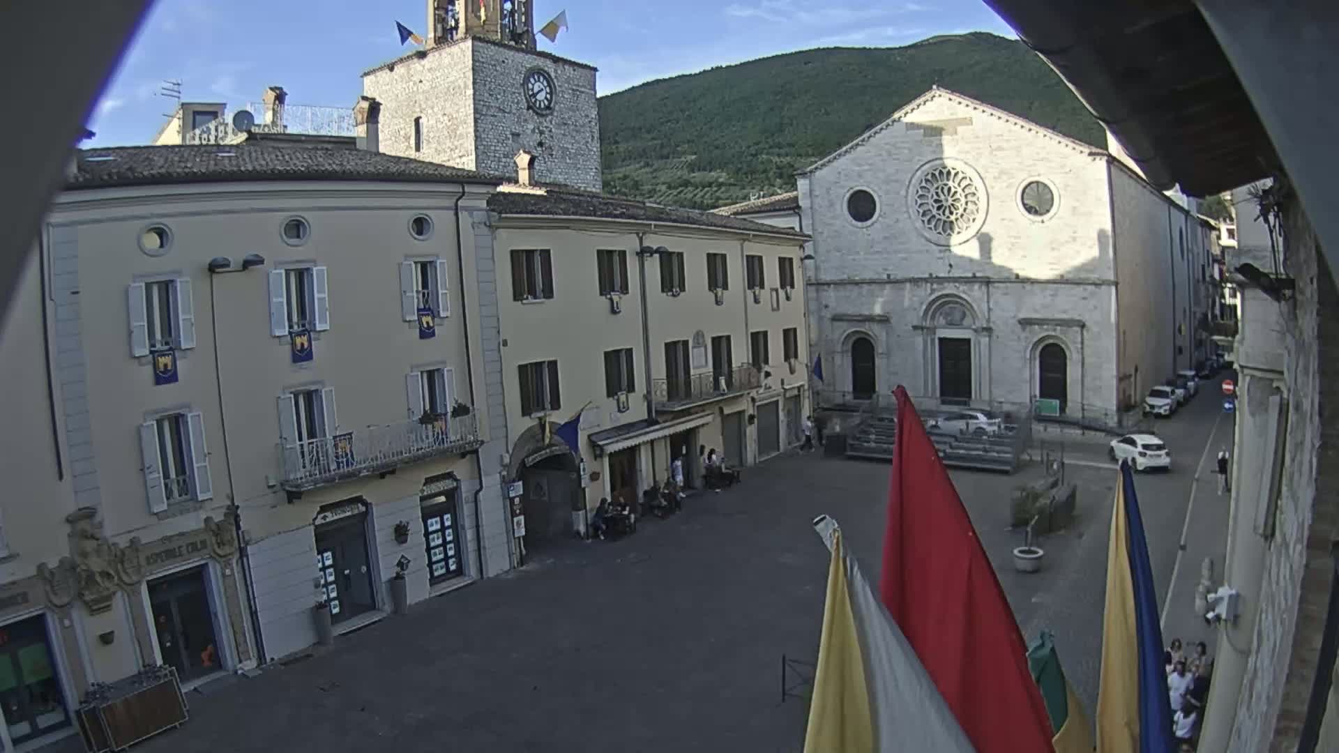 A town square with several multi-story buildings, including a church with a rose window, under a mostly clear sky with a mountain in the background.