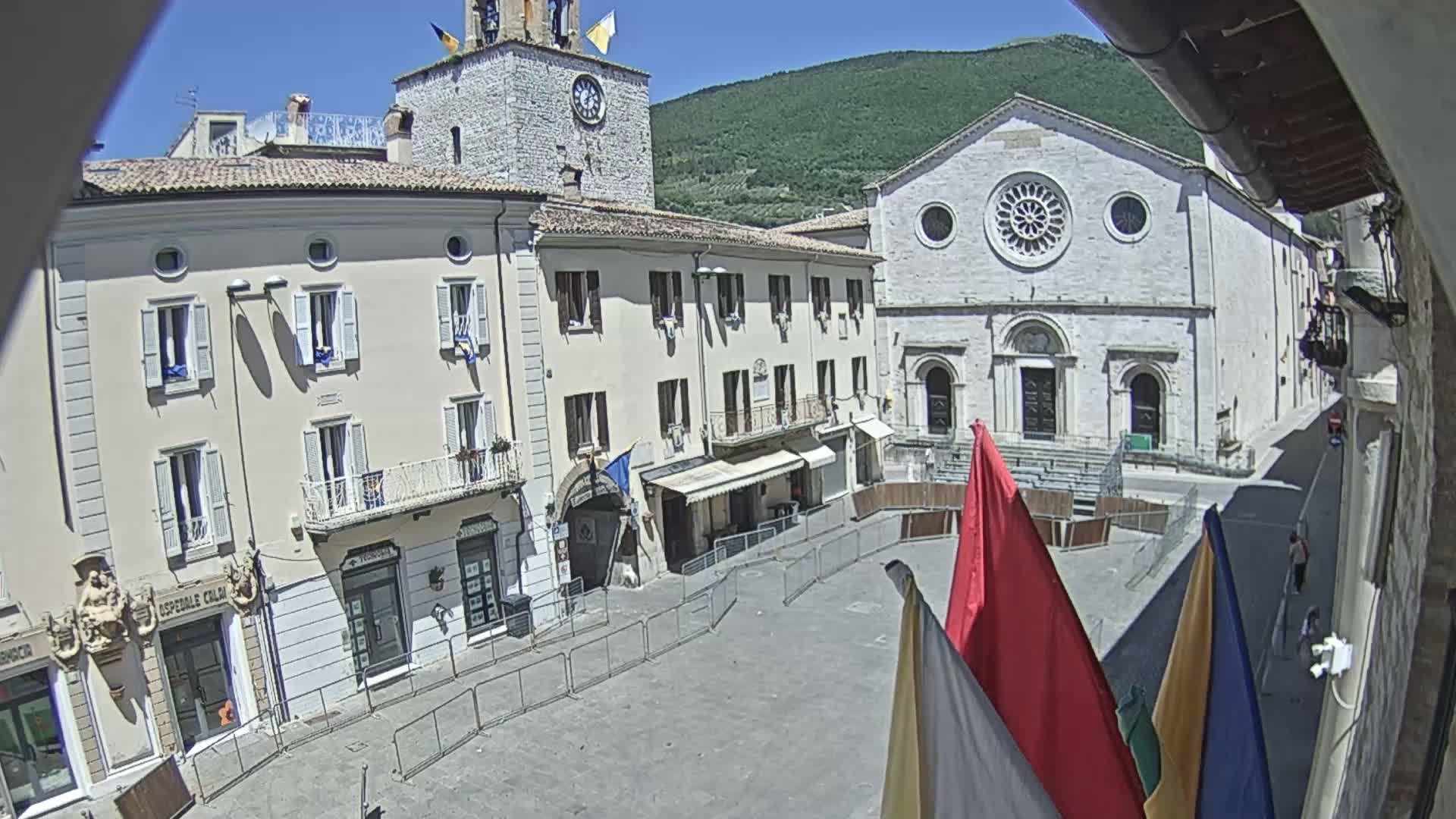 A sunny day reveals a town square, partially enclosed by metal barriers, with several buildings including a church with a rose window and a clock tower, set against a backdrop of green hills.