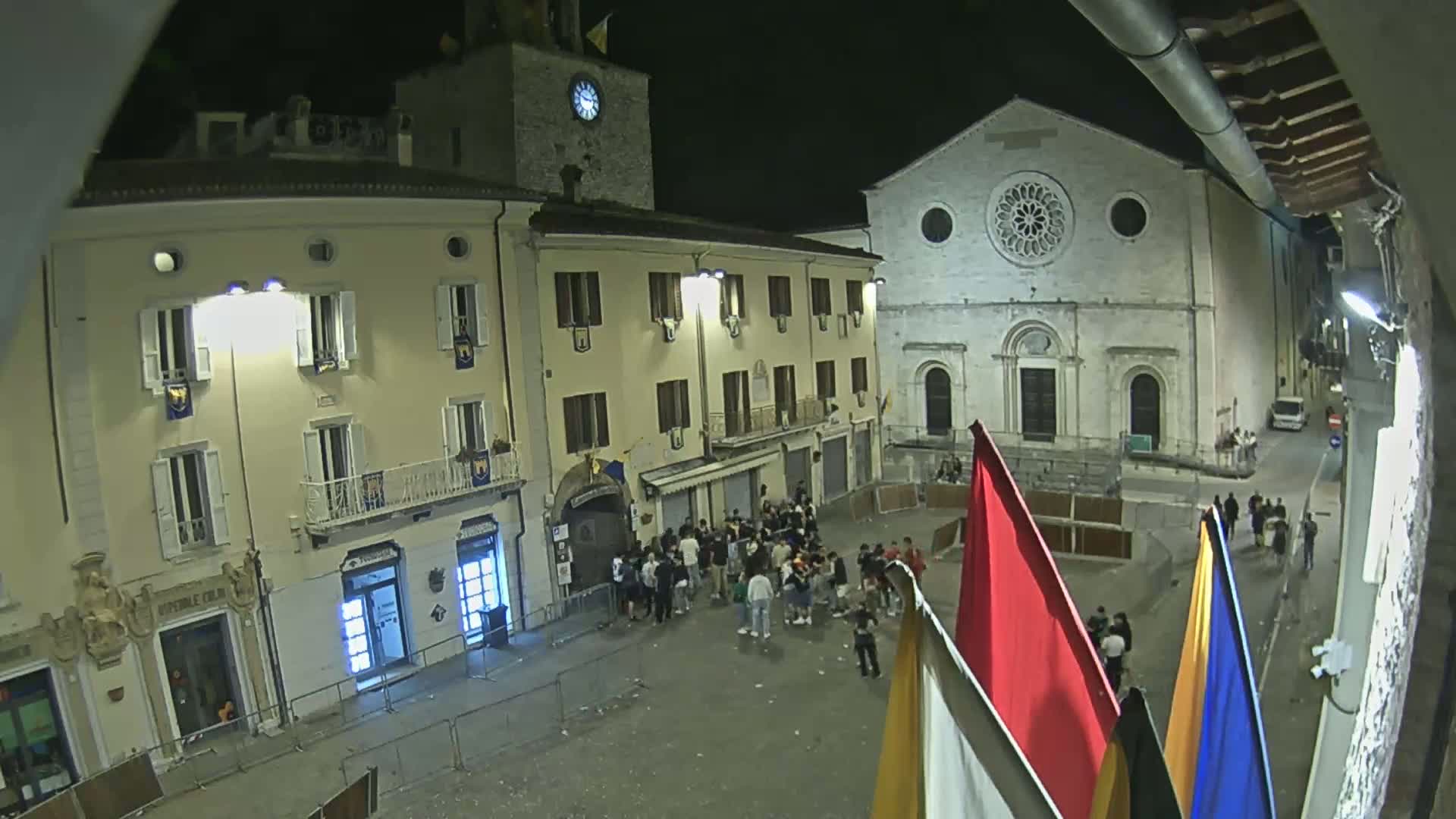 A nighttime view of a town square with several buildings, including a church with a rose window, and a crowd of people gathered in the center, under clear skies.