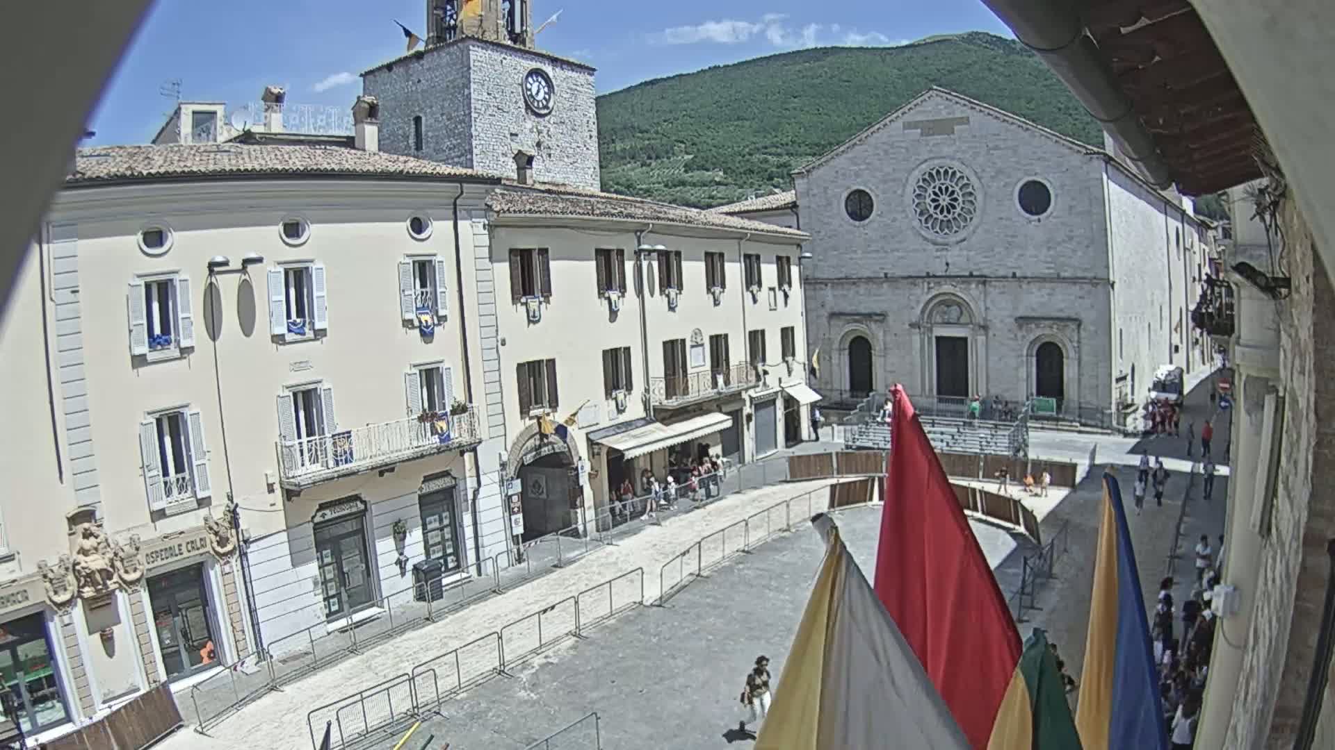 A sunny town square is partially enclosed by temporary fencing, with several buildings, including a church with a rose window, visible in the background.