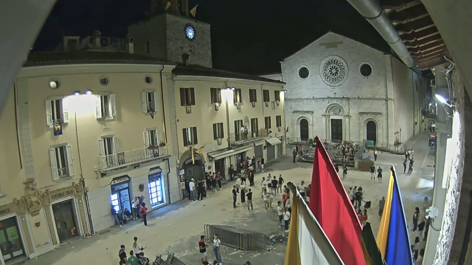 A nighttime, outdoor town square scene shows several people gathered in front of buildings, including a church with a large rose window, under what appears to be clear skies.
