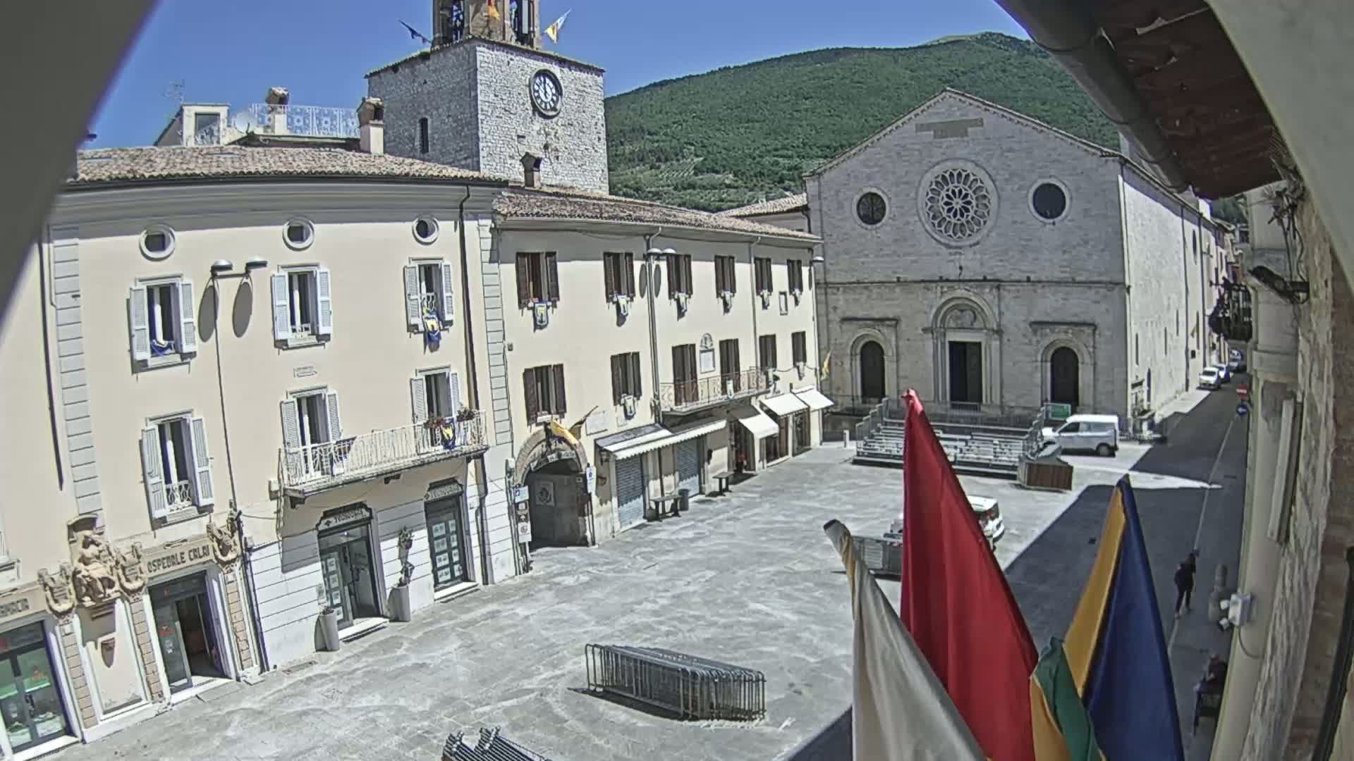 A town square in a sunny, mountainous region is surrounded by light-colored buildings, including a church with a rose window, and a clock tower.